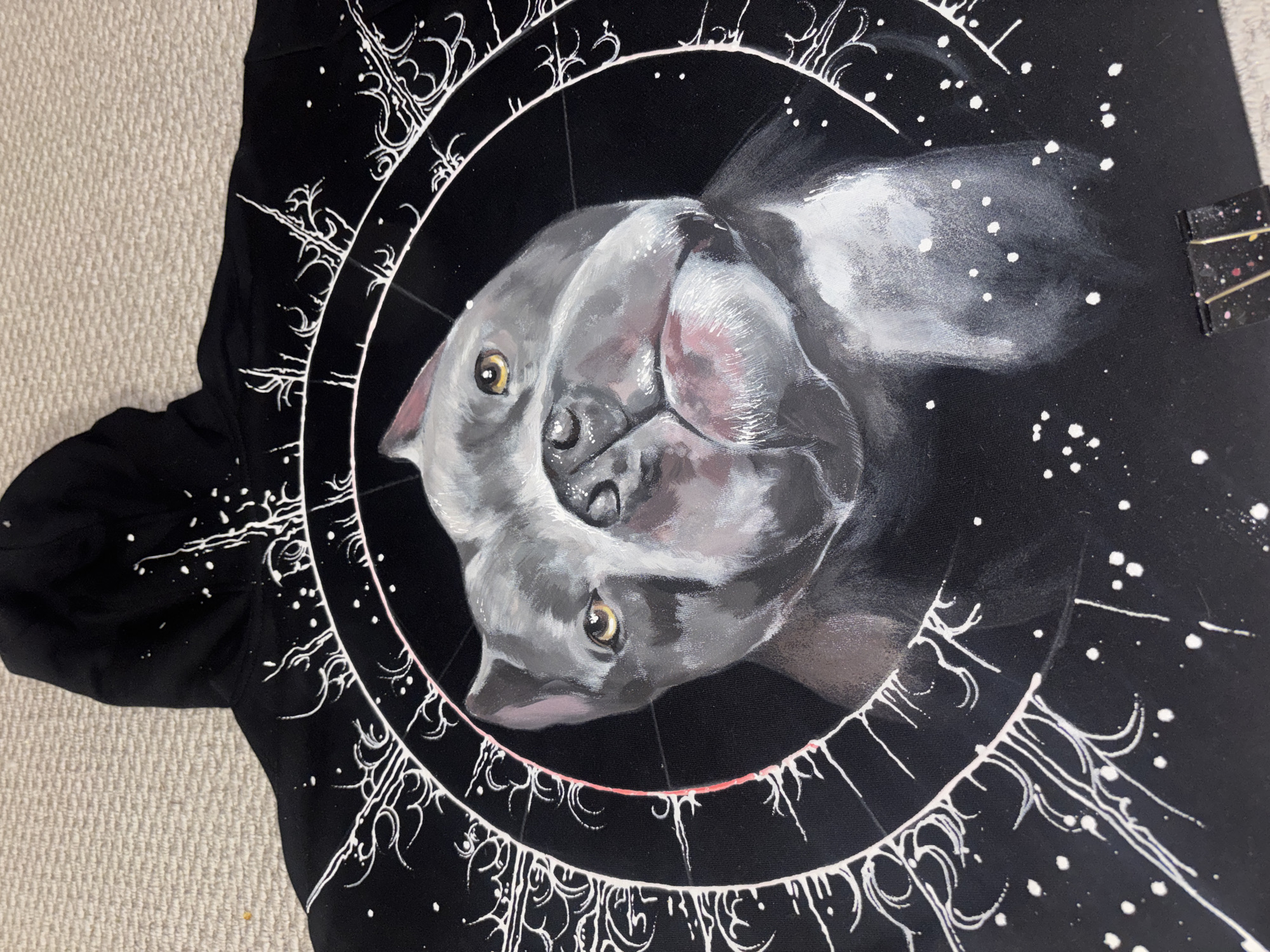 A black hoodie with a painted design is lying on a carpeted surface. The design features a gray dog lying on its back within a white circular border. The border is adorned with white geometric patterns and dots, resembling celestial elements. To the right of the design is a small black object with gold lines and dots on it. The hoodie's sleeve is visible on the left side of the image. The painting is likely acrylic. There are no signs indicating this is related to Krasnoyarsk, Russia.