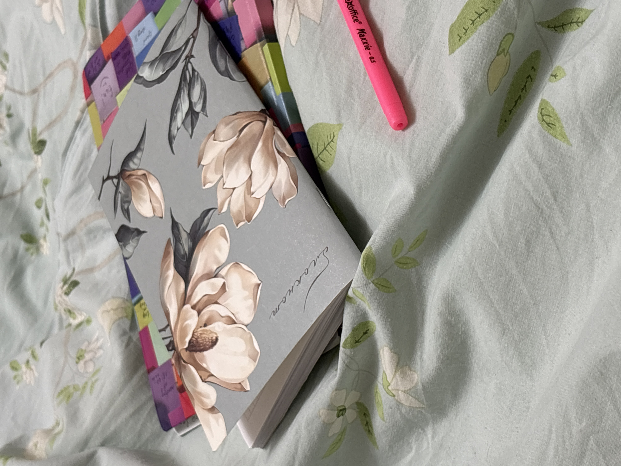 The image shows a bed covered in a light blue floral patterned sheet. On the bed, there is a closed notebook with a gray cover and magnolia flower design. The notebook has colored tabs sticking out from its side. A pink marker is lying on the bed near the notebook. The location of the scene is not possible to be determined based on the information provided.