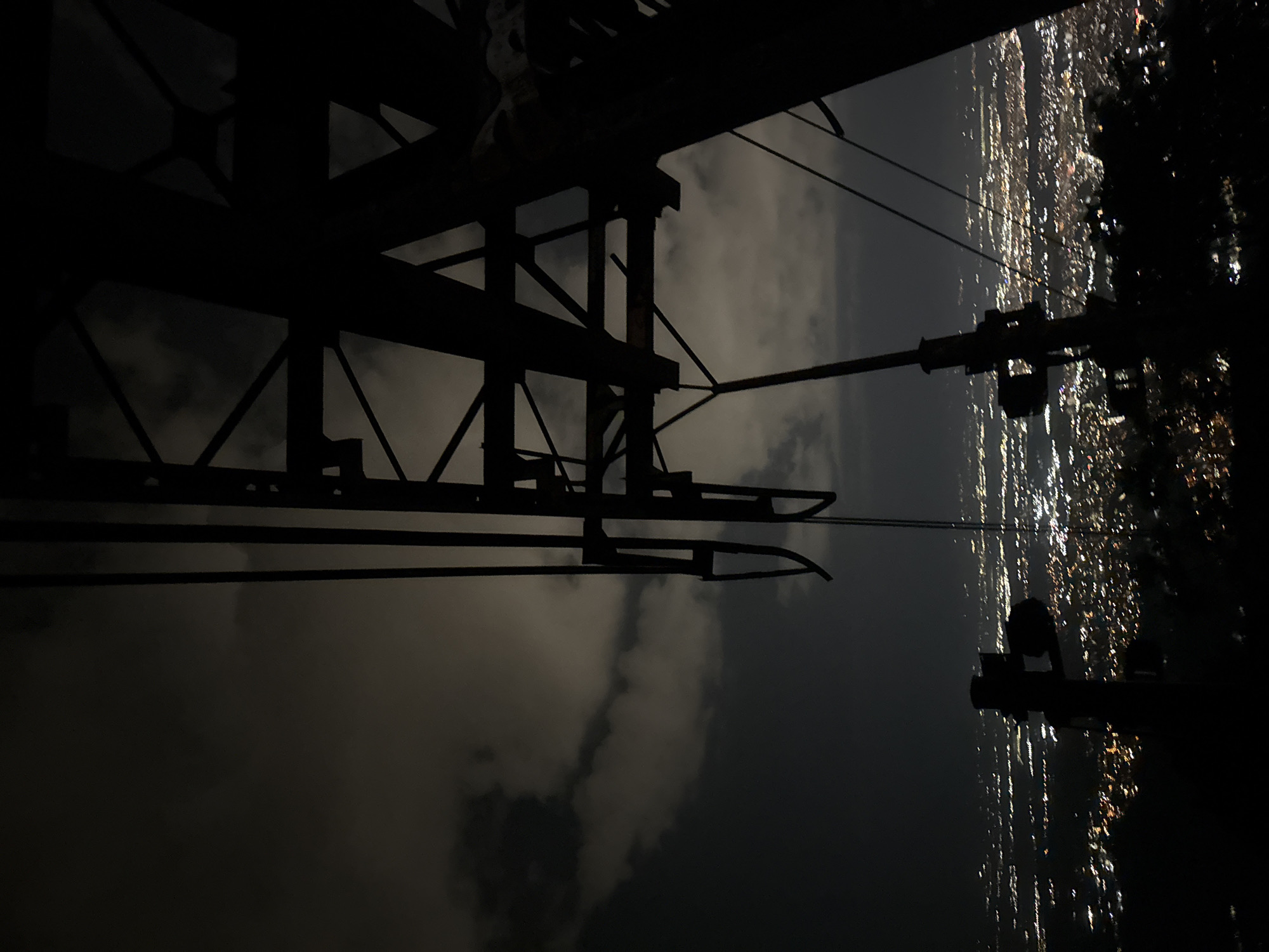 The image presents a detailed nighttime view of an urban landscape from an elevated perspective. In the immediate foreground, a large, dark silhouette of a metallic cable car pylon structure is prominently positioned, extending from the right side of the frame and upward, with visible vertical supports, diagonal bracing, and multiple cables extending downwards. Below this structure, the city of Sofia, Bulgaria, stretches into the distance, densely illuminated by a myriad of artificial lights that form a blurred, sparkling pattern across the terrain. The sky above is dark, featuring numerous irregularly shaped clouds that are subtly illuminated from below by the city's light pollution. No individuals or specific actions are discernible within the frame. The scene is consistent with a vantage point from Vitosha Mountain, overlooking the city of Sofia.