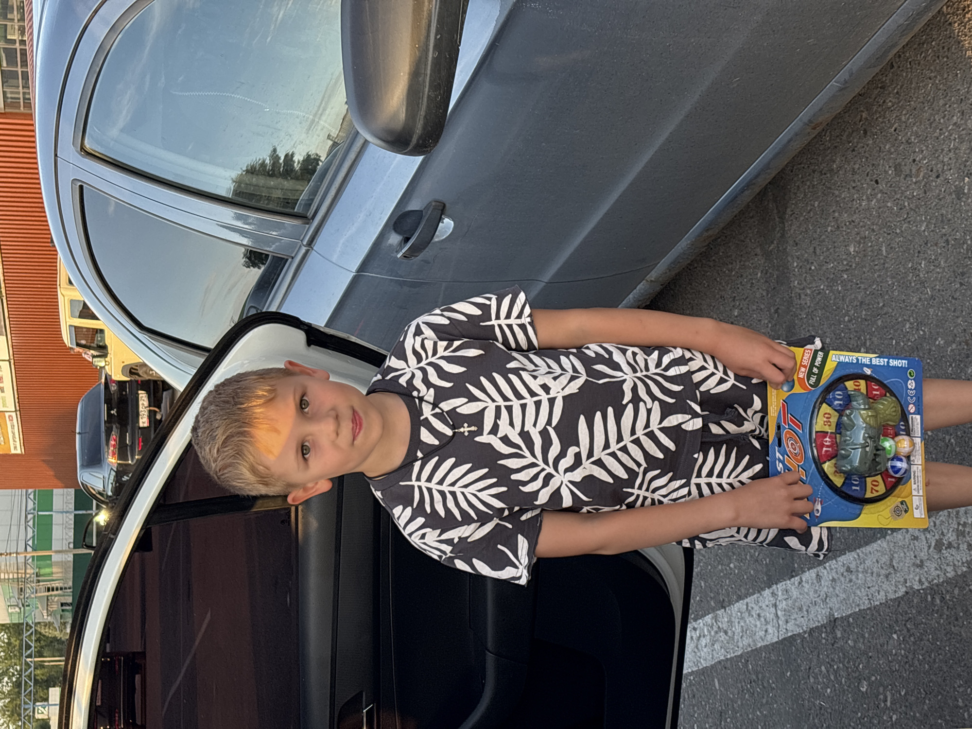 A boy stands in front of a car holding a toy. He has blond hair and is wearing a dark t-shirt with a white leaf pattern and a small silver cross necklace. The car is a light blue sedan with its doors open. The boy is holding a toy target game with a blue and yellow package. The background shows a building with an orange corrugated metal wall and a parking lot. The lighting suggests it is late afternoon.