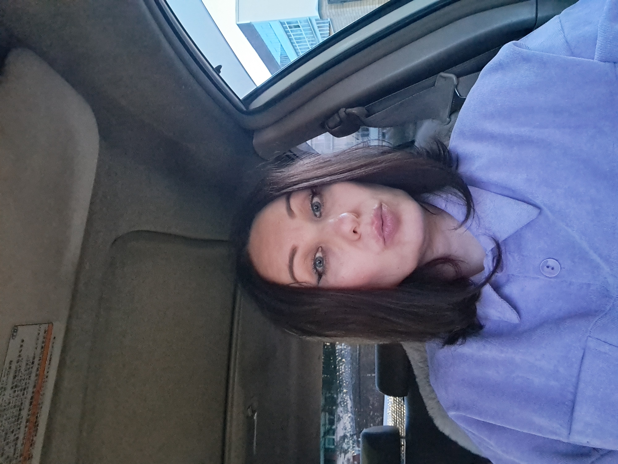 A woman with dark hair and winged eyeliner is sitting in the driver's seat of a car, looking towards the camera with her lips pursed. She is wearing a light purple corduroy shirt. The interior of the car, including the headliner and a car seat, is visible. Through the passenger side window, a modern building with blue and white accents can be seen, suggesting an urban environment. A sticker with Japanese text is visible on the sun visor. The lighting suggests it is daytime.