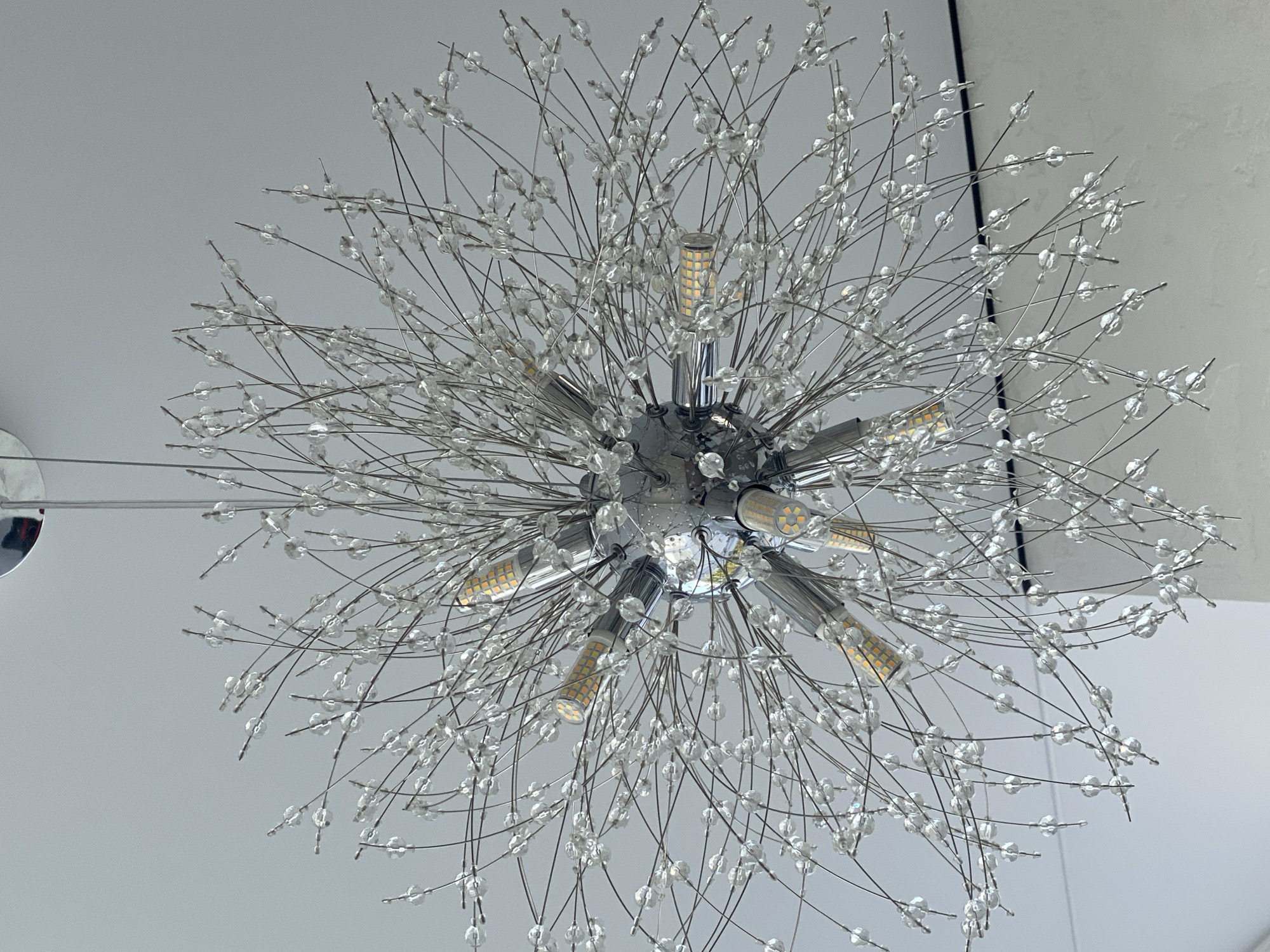 A close-up, low-angle shot shows a modern, spherical chandelier hanging from a white ceiling. The chandelier is made of a central chrome sphere from which numerous thin metal wires extend outwards, forming a dandelion-like shape. Each wire is adorned with small, clear, faceted beads, giving the impression of a glittering explosion. Several small, cylindrical LED light bulbs are interspersed among the wires, emitting a warm, yellowish light. The background is a plain white ceiling. The location is unknown.