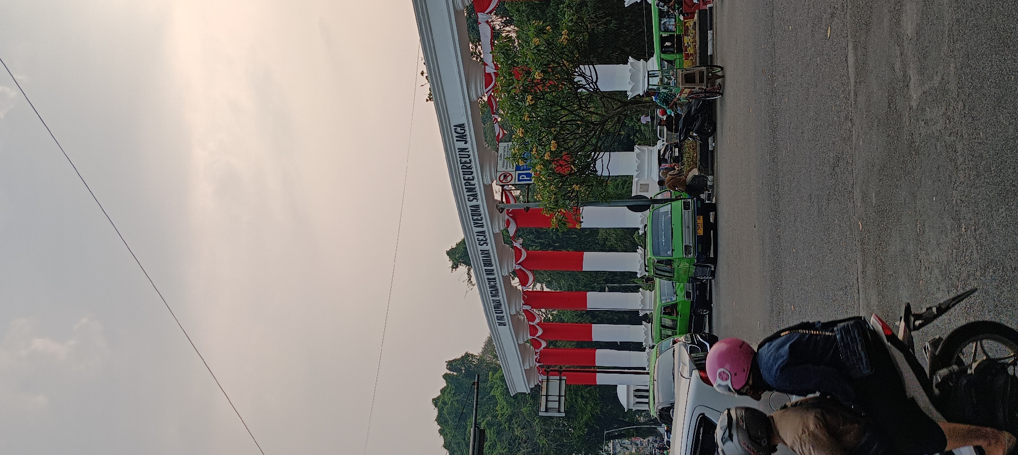 In Bogor, Indonesia, a street scene unfolds at what appears to be a significant gateway. The gateway, decorated with red and white stripes alternating with white pillars, bears an inscription that reads "DI NU KIWARI NGANCIK NU BIHARI SEJA AYEUNA SAMPEUREUN JAGA". This text is likely in Sundanese, a language spoken in West Java, hinting at the location's cultural context.
The street is busy with traffic. Several green minibusses, a common mode of public transport in Indonesia, are visible. One of them has a license plate starting with "F," which is the code for Bogor Regency and City. A white sedan is also present, and in the foreground, two people are riding a motorcycle. The person in the back is wearing a pink helmet.
Other elements in the scene include a traffic light, a parking sign, and a vendor pushing a cart. Lush green trees and foliage frame the background, suggesting a well-treed urban environment. The sky is overcast with a hint of a sunset glow, indicating it is late afternoon or early evening. The overall impression is a snapshot of everyday life in a bustling Indonesian city, marked by distinctive local architecture and transportation.