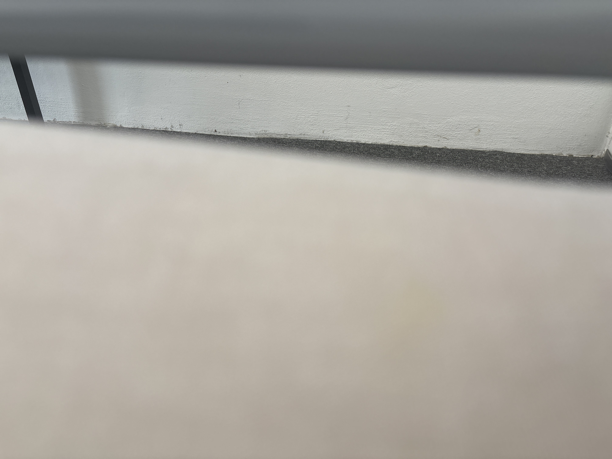 The image is an indoor shot, focusing on the lower portion of a wall and the floor. The wall is a textured, off-white color, with a darker, granular floor meeting it at the base. Above the wall, a dark, smooth horizontal surface is visible, casting a shadow on the wall. To the left, a black vertical object casts a diagonal shadow on the wall and floor. The foreground is dominated by a soft, out-of-focus, light-colored surface, possibly a piece of furniture or another wall, taking up the lower half of the frame. There are no people or animals present. The scene appears to be in a common indoor space, such as an office or a hallway.