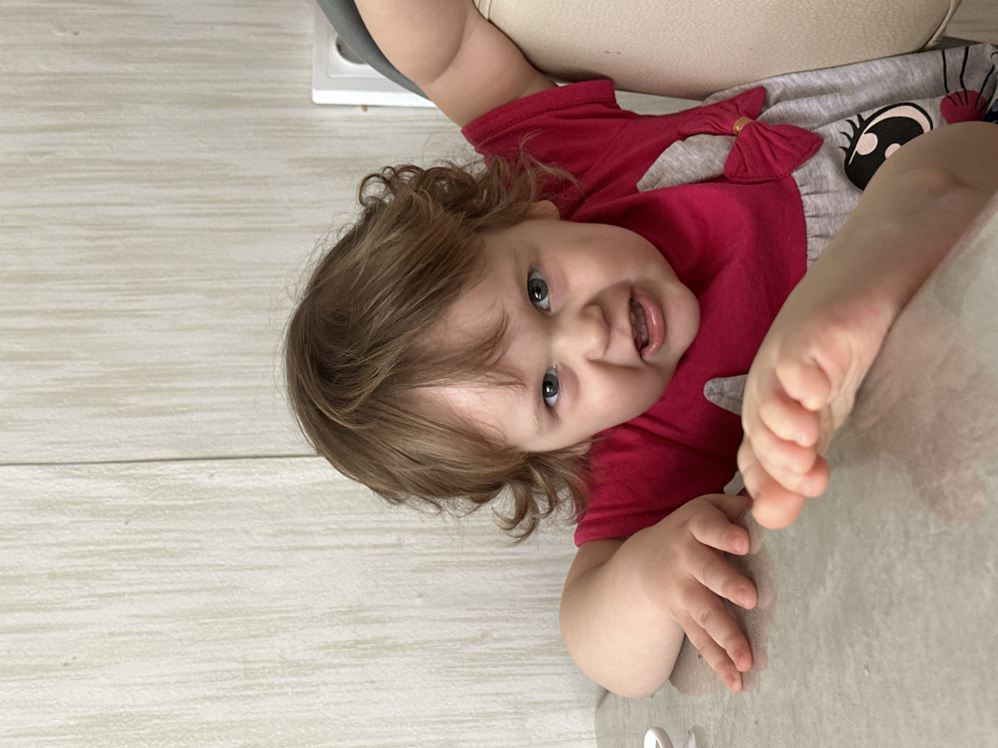 A baby with curly brown hair and blue eyes lies on a light-colored surface. The baby wears a red shirt with a grey top featuring a cartoon eye graphic. The baby's feet are close to the camera. The surface appears to be a table or counter with a light-colored, possibly wooden, floor in the background. The location is unspecified; the statement that the scene is in Chaikovsky, Russia, cannot be confirmed from the image.