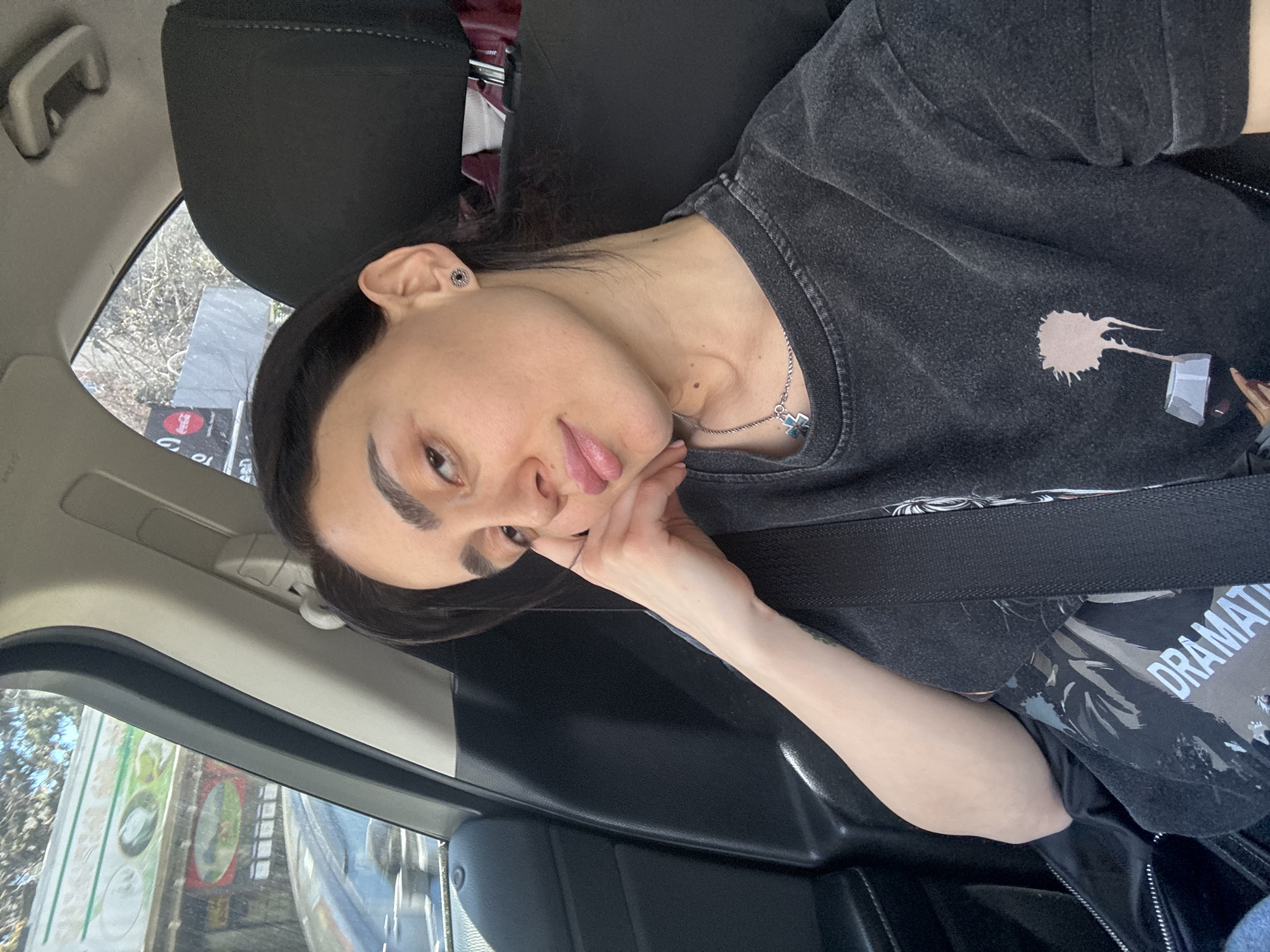 A young woman is seated in the passenger seat of a car, possibly in Tbilisi, Georgia. She is wearing a black t-shirt with a graphic print, a seatbelt, and a silver necklace. Her face is turned toward the camera, and she rests her chin on her hand. The car appears to be parked on a street with storefronts visible through the window.