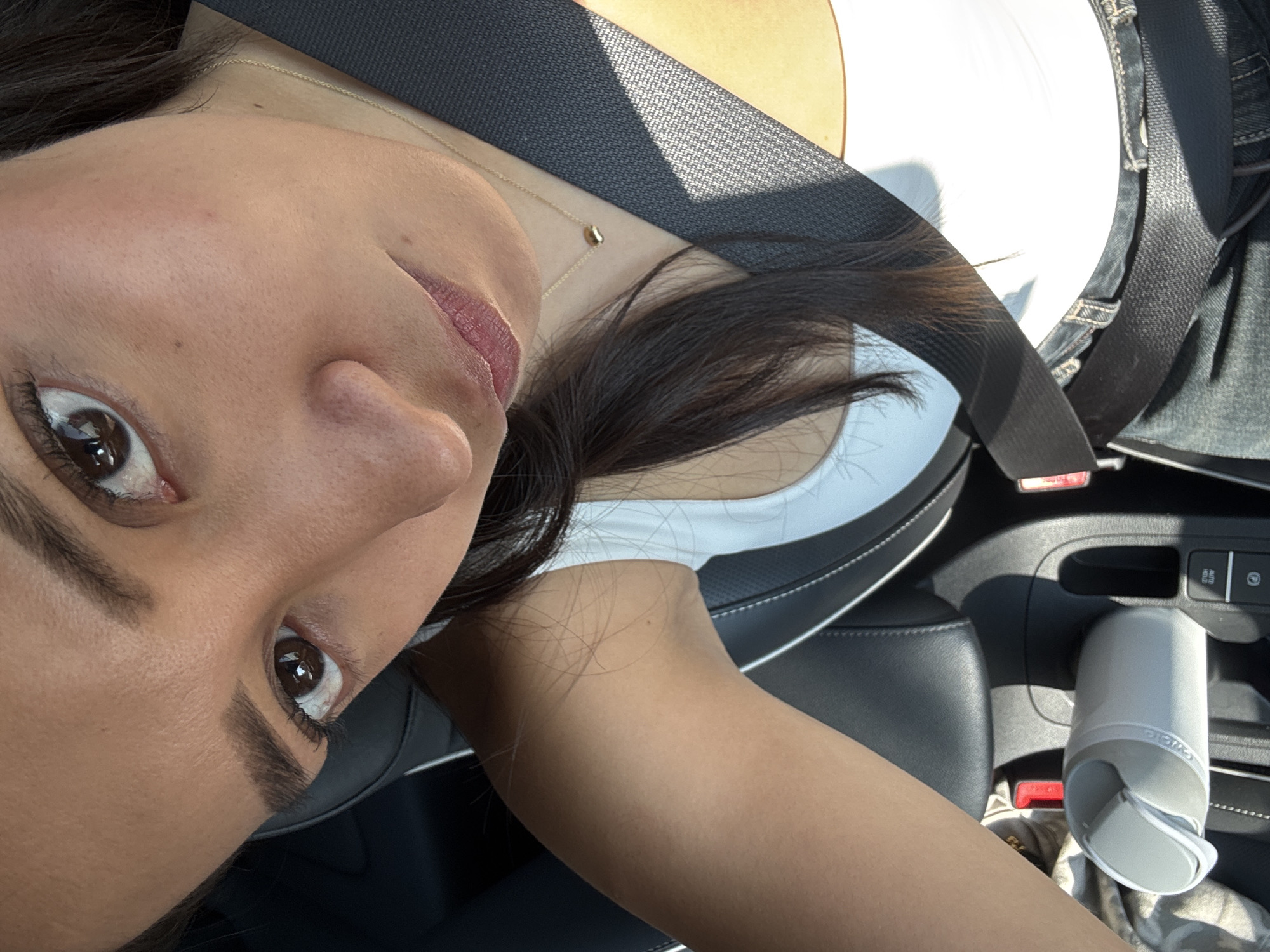A close-up, tilted, eye-level shot depicts a woman wearing a seatbelt in a car. Her face is the primary focus, showing brown eyes, dark eyebrows, and pink-tinted lips. She also wears a gold necklace. The background shows the interior of a car: a white tank top beneath the seatbelt, dark gray seat, and a cup holder with a gray water bottle. Specific city or location is not discernible from the details in the image.