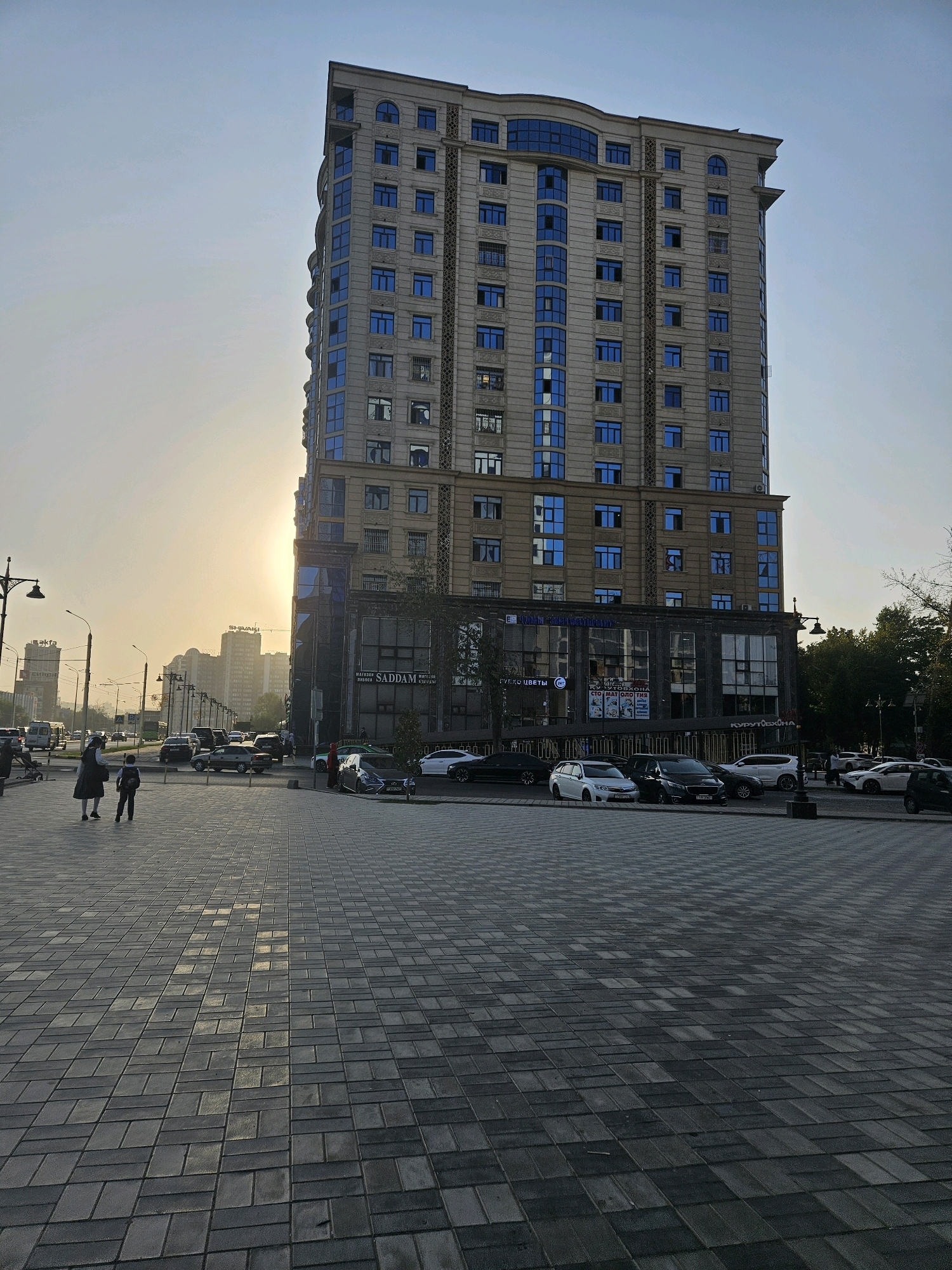 The image shows a modern, tall building in Dushanbe, Tajikistan, with a clear sky and sun low on the horizon. The building, likely an office or residential complex, features light-colored stone cladding and numerous blue-tinted windows. The ground level of the building appears to house commercial establishments, with signage visible, including one that reads "SADDAM". In the foreground, a paved pedestrian walkway is visible, with two individuals, a woman and a child, walking away from the camera. Several cars are parked along the street in front of the building and also on the opposite side of the street. The overall scene depicts an urban environment during what appears to be either sunrise or sunset, with a slightly hazy atmosphere. The architecture suggests a contemporary style prevalent in urban centers.