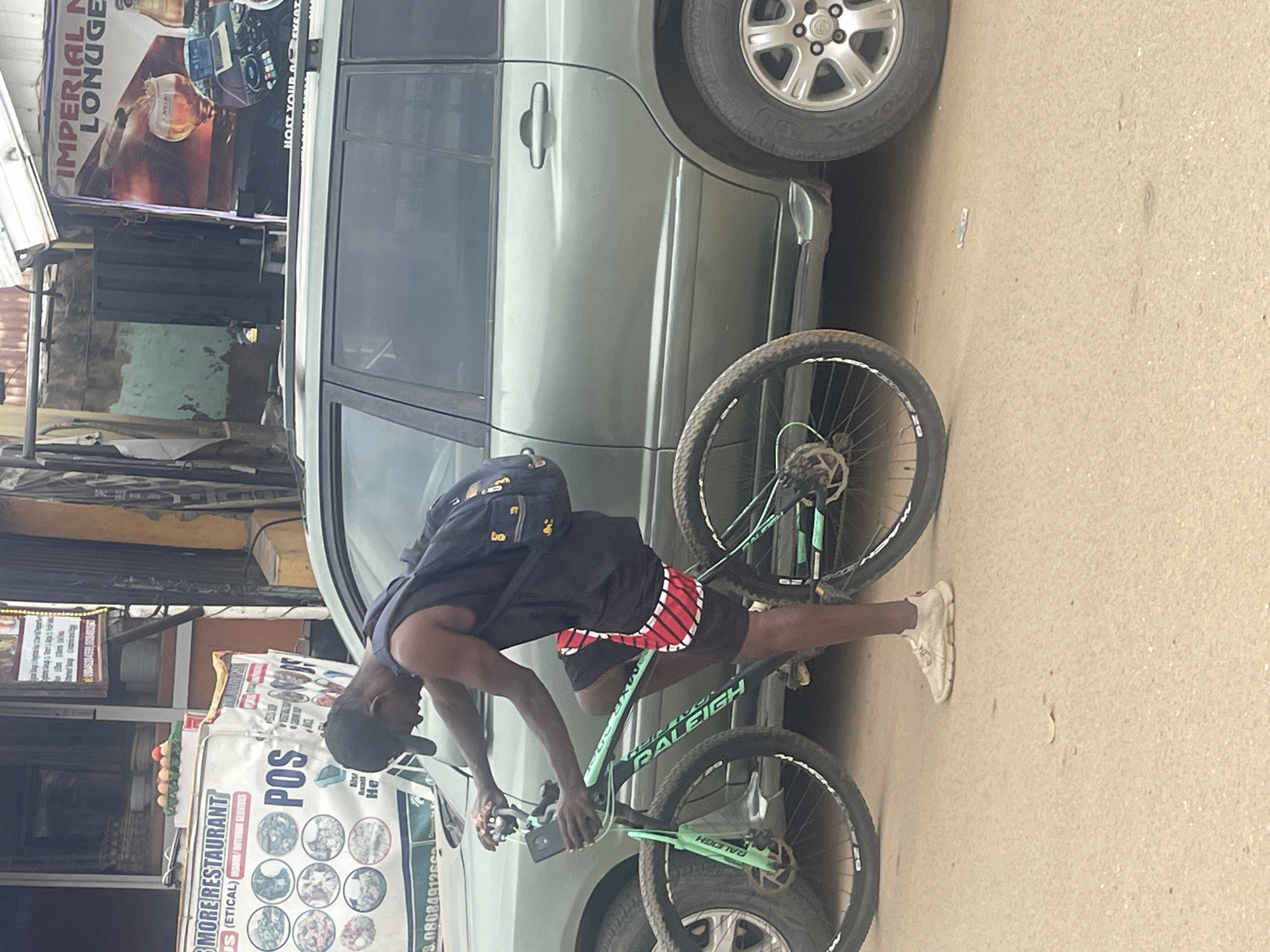 This media file shows a young man wearing a dark blue t-shirt, black shorts with red and white stripes, and white sneakers, leaning over a light green Raleigh bicycle. He has a dark backpack on his back and is looking down at his phone, which he is holding with his left hand. The bicycle is parked next to a light green vehicle, possibly an SUV, with its door open.
In the background, there are several signs and posters. One poster advertises "MORE RESTAURANT" and mentions "US (ETICAL) DOOR/OUTDOOR SERVICES" with a phone number. Another sign has the text "POS" and shows various food images. A large banner displays "IMPERIAL M LONUGE". The scene appears to be on a street in Abuja, Nigeria, given the location context. The lighting suggests it could be daytime, possibly overcast or late afternoon, as there are no harsh shadows. The man's posture and focus on his phone suggest he is engaged in a personal activity. The overall scene captures a slice of everyday life in an urban setting.