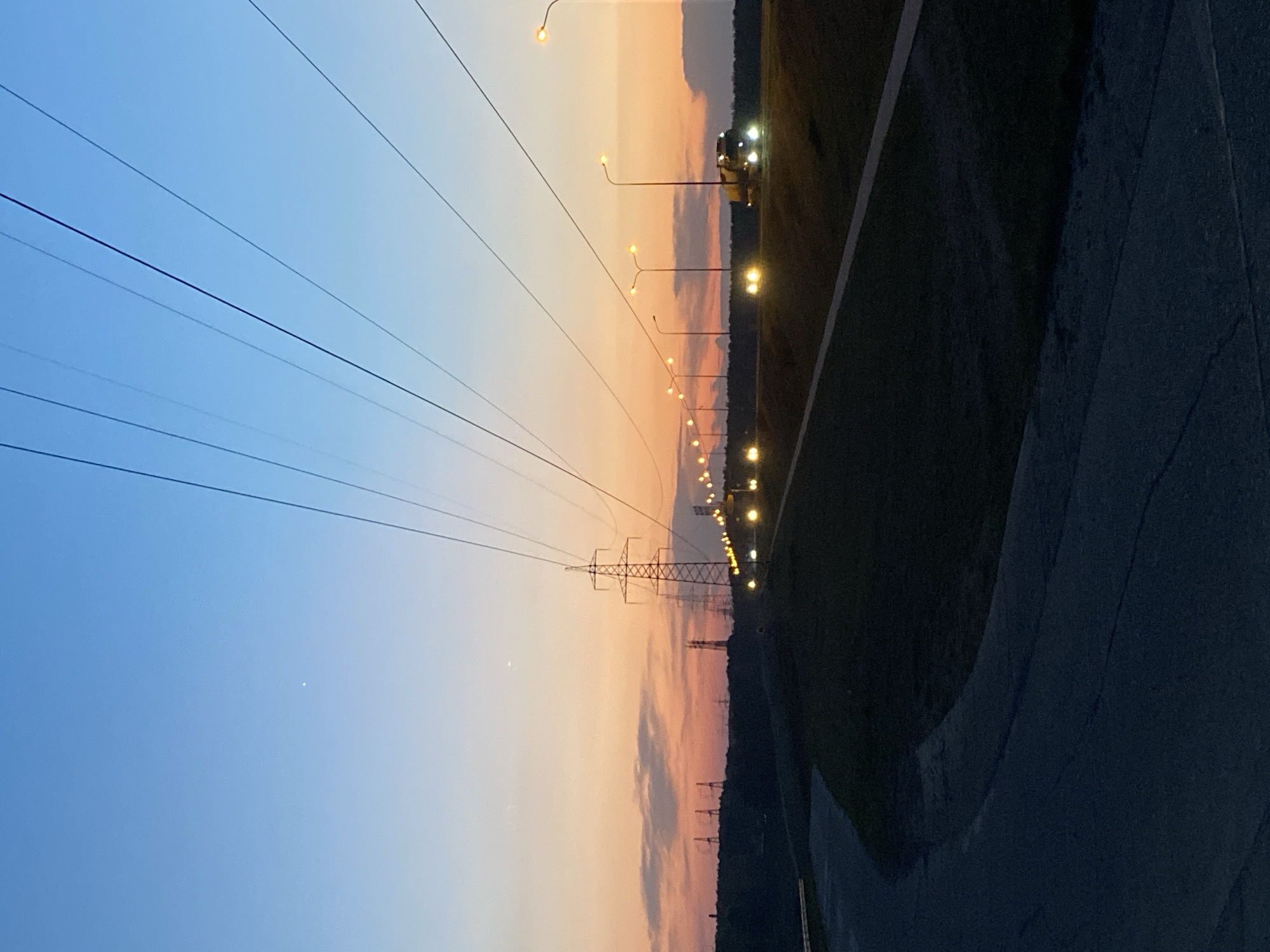 This photograph shows a twilight view in what appears to be an urban or suburban setting. The scene includes power lines running across the sky, supported by a tall, lattice-structured transmission tower. Alongside the power lines are street lights with glowing bulbs, suggesting it is dusk or dawn. In the distance, there are additional lights and what seems to be a grassy area bordering a road. The road is dark and stretches into the foreground. The sky transitions in color, from a blue hue to a warm, orange glow, indicative of the setting or rising sun. Given the context of the city of Chaikovsky, Russia, it is reasonable to assume that the landscape features are related to its local environment and infrastructure. There are no people visible in the photograph.