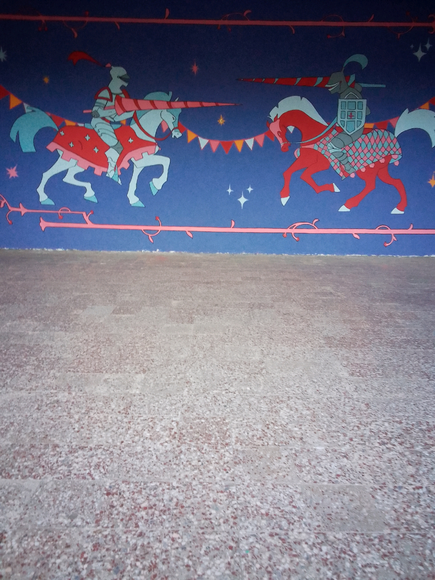 The image shows a section of a wall mural depicting a jousting scene. Two knights on horseback, rendered in a stylized, cartoonish manner with bold outlines and flat colors, are the central subjects. The knight on the left rides a light blue horse with red barding and a red lance, while the knight on the right rides a red horse with a checkered blue and pink blanket and a grey shield. Both knights are clad in armor. The background is a deep blue, adorned with small yellow and white stars. Decorative elements in pink, red, and yellow, resembling banners and vines, frame the scene at the top and bottom. The bottom portion of the image shows a textured, mottled floor, possibly concrete or terrazzo, with flecks of red, white, and grey. The mural appears to be on an interior wall, possibly in a public space. The setting is not specified as being in Tbilisi, Georgia, and no specific landmarks or identifying features of that city are visible.