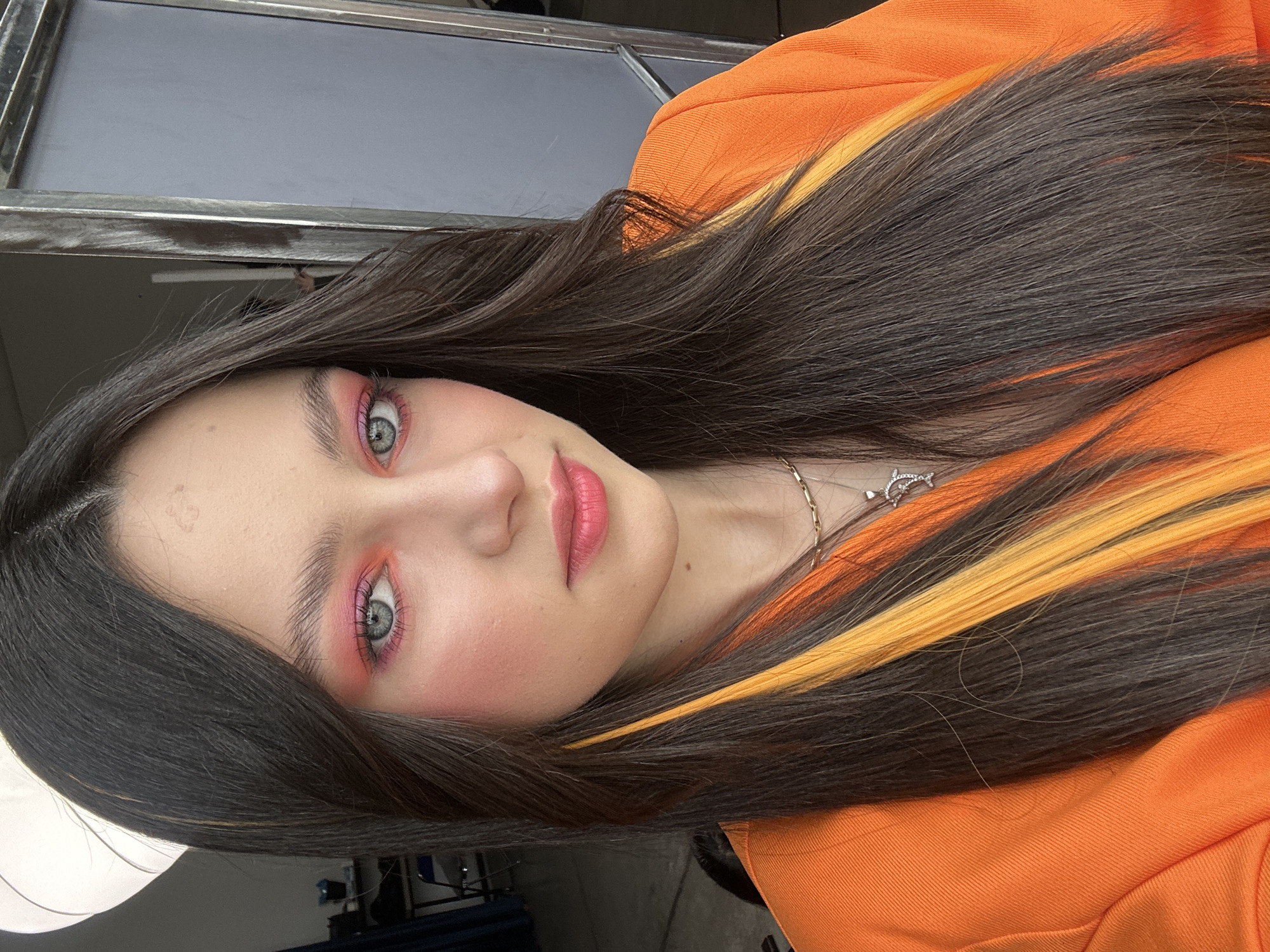 A close-up, eye-level, rotated portrait of a fair-skinned young woman with long, dark brown hair highlighted with streaks of yellow-orange. She has pink blush and matching eye shadow. The environment is a film set, as suggested by equipment in the background. No information suggests the city of Yerevan, Armenia.
