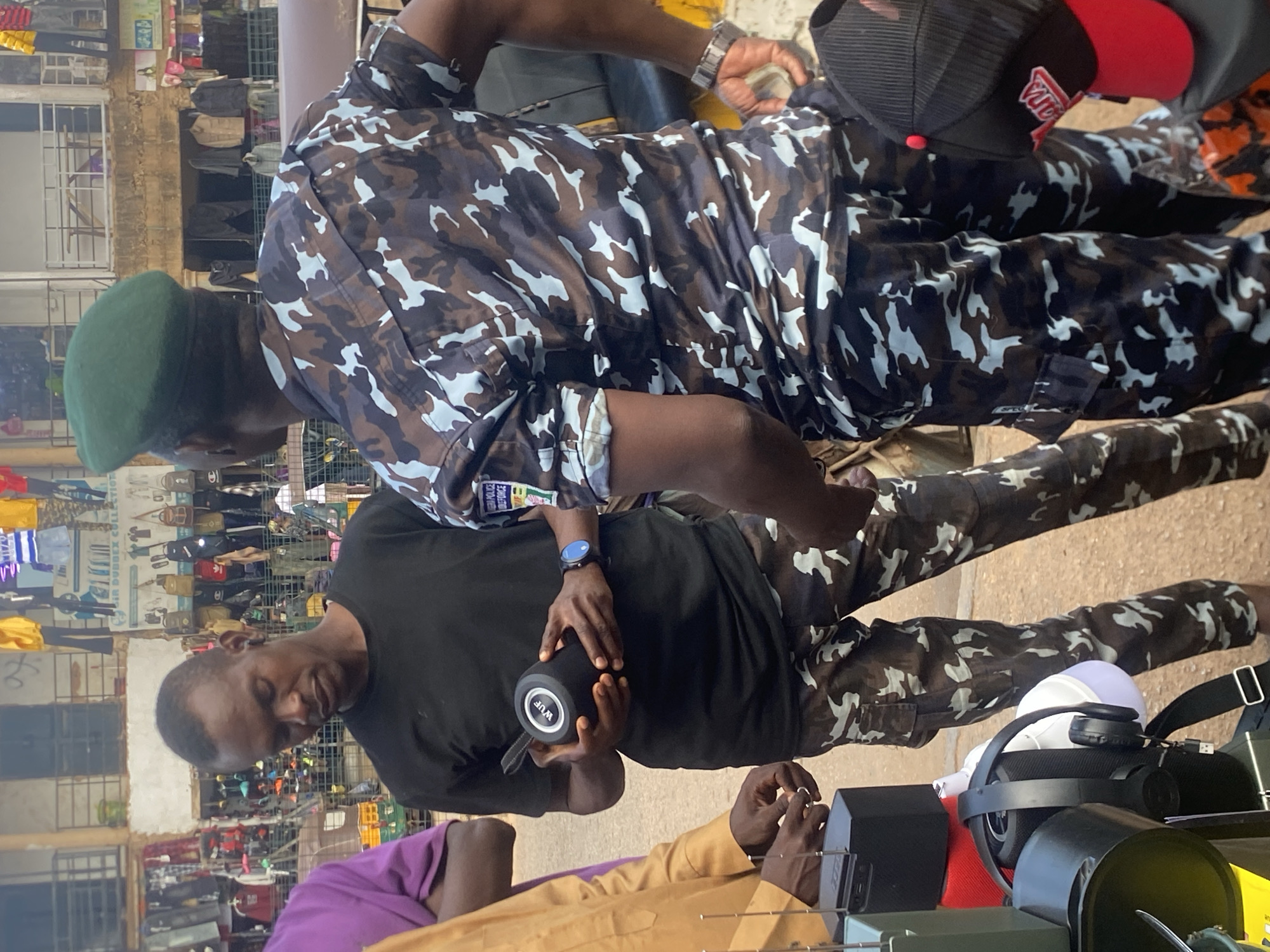 In Bukuru, Nigeria, a man wearing a black t-shirt and camouflage pants holds a black cylindrical speaker with a white "WUF" logo. He is standing near a person in a camouflage police uniform, distinguished by a green beret and a blue, black, and white camouflage pattern on their shirt. The police officer's shirt displays a Nigerian Police Force emblem on the sleeve. Other individuals, one in a purple garment and another in a yellow garment, are partially visible. The background reveals a bustling market environment with stalls displaying clothing and accessories. In the foreground, various electronic devices, including a black speaker, headphones, and a white illuminated object, are arranged on a surface. The scene appears to be an outdoor transaction or interaction within a marketplace setting.