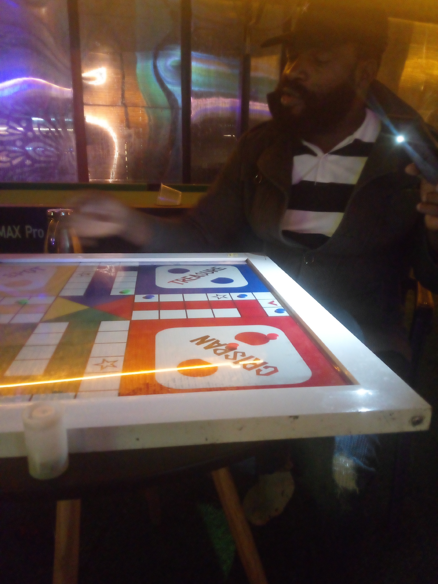 In a dimly lit room, a man with dark skin and a beard, wearing a black jacket over a black and white striped polo shirt, is seated at a table. He is looking towards the right side of the frame, with a slight glow on his face from a nearby light source, possibly a phone he is holding. In front of him is a Ludo board game, brightly colored with red, blue, purple, and orange sections, featuring white squares and stylized stars. The board has text that is partially visible, including "TREASURE" and "GRISPAN". The background is out of focus, showing reflections of colorful lights on a textured surface, suggesting a vibrant or perhaps a commercial environment, potentially a bar or entertainment venue in Barakin Lerawi, Nigeria. A small, translucent container is on the left side of the table. The overall atmosphere is informal and suggests a moment of leisure or friendly competition.