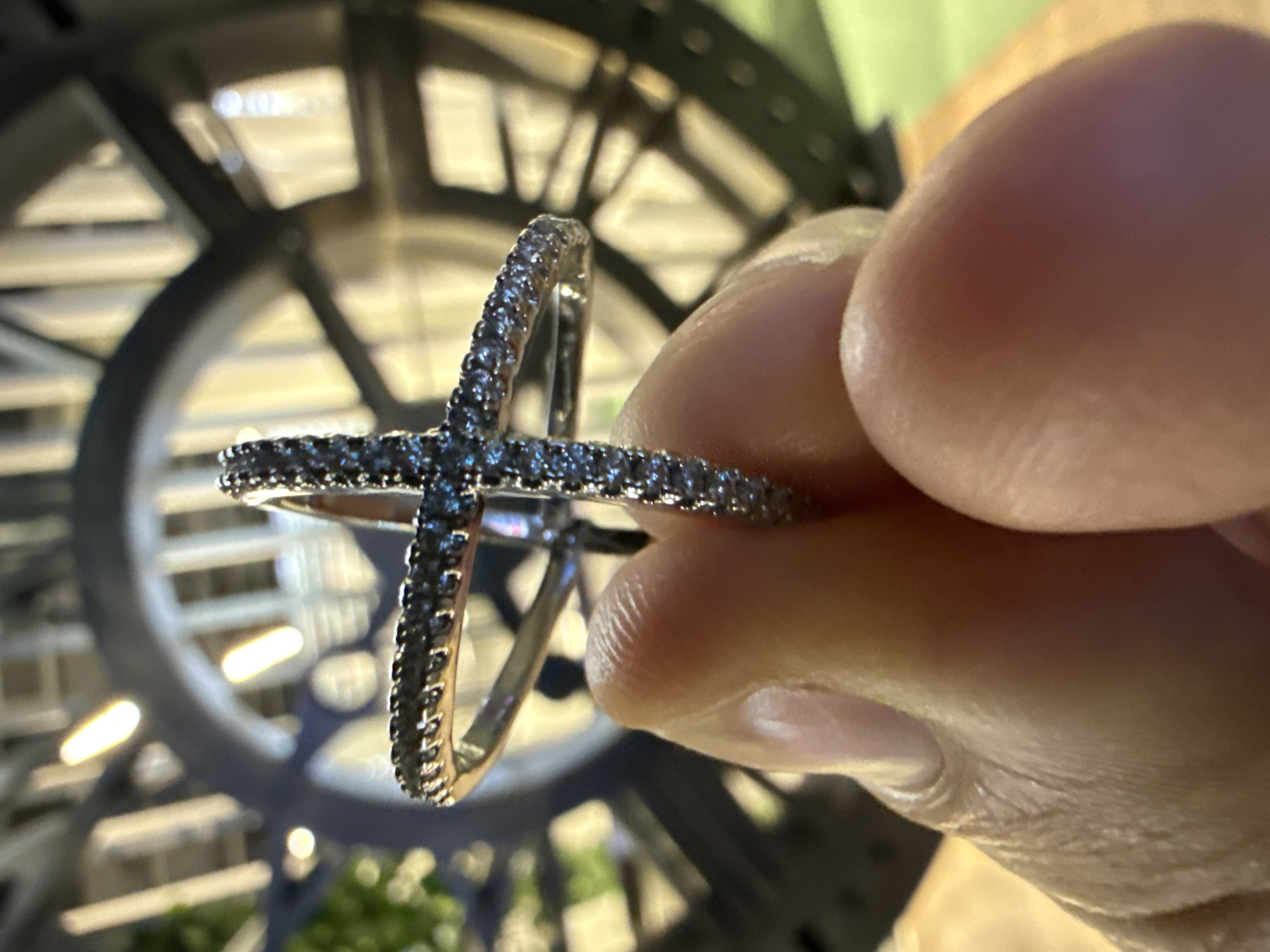 A person holds a silver-colored ring with dark stones against a backdrop of a large, circular, metallic structure. The structure has a radial design with crossbeams and circular supports, creating a complex pattern. The ring, held between the person's fingers, is formed by two crossing bands, each studded with small, dark stones. The skin of the hand appears light-toned, with visible fingerprints and folds. The background is blurred, but hints of greenery are visible beneath the metallic structure. This image was taken in Moscow, Russia, as the structure in the background is Moscow's Central Children's Store clock.