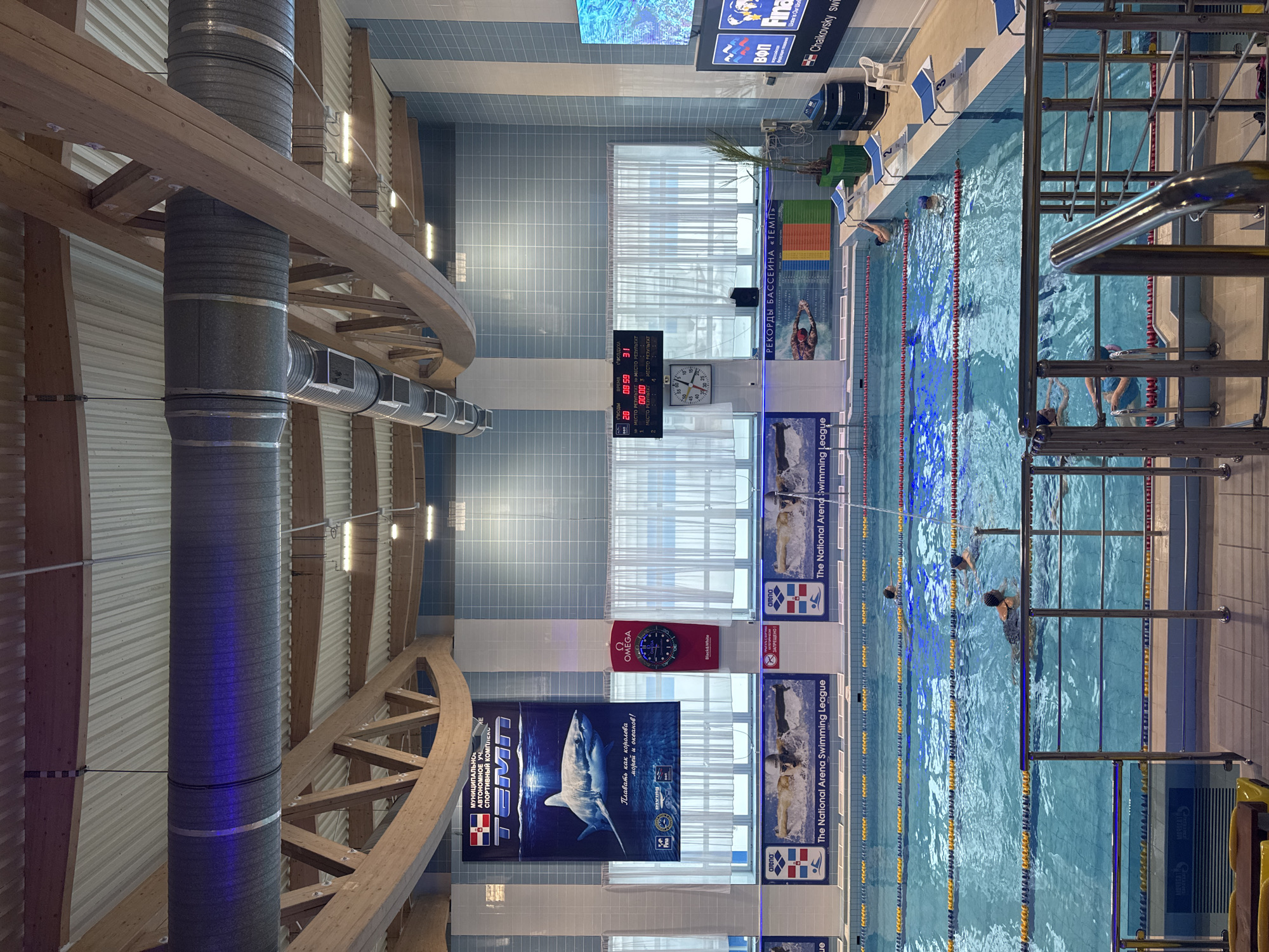 The image depicts an indoor swimming pool facility, specifically the TEMPO Sports Complex in Chaikovsky, Russia. The scene is brightly lit, showcasing a multi-lane pool with turquoise water, marked by yellow, blue, and red lane lines. Approximately 6-8 individuals, appearing to be adults or adolescents, are present in the water, engaged in swimming or standing. One person is observed climbing out of the pool near starting block number 6, while others swim across various lanes.
The facility features a high ceiling with exposed wooden truss beams, corrugated metal roofing, and large grey cylindrical HVAC ducts running across, some illuminated with blue light accents. The walls are tiled in alternating light blue and white vertical stripes. Along the back wall, large windows are covered by white horizontal blinds. Prominent signage includes a large banner on the left displaying a great white shark with Russian text "ТЕМП" (TEMPO) and an Omega brand clock/display below it. Multiple smaller banners along the pool's edge advertise "The National Arena Swimming League." A digital scoreboard on the back wall shows "20," "09:59," and "31," with an analog clock mounted beneath it. Another sign displays "РЕКОРДЫ БАССЕЙНА «ТЕМП»" (Pool Records "TEMPO"). On the far right, a large digital screen shows an underwater image and text including "Final" and "Chaikovsky swimming." Starting blocks are visible on the right side of the pool, numbered 1 through 6. The deck area features white plastic chairs and two large potted plants. A polished metal railing system is present in the foreground, indicating a viewing or access area.