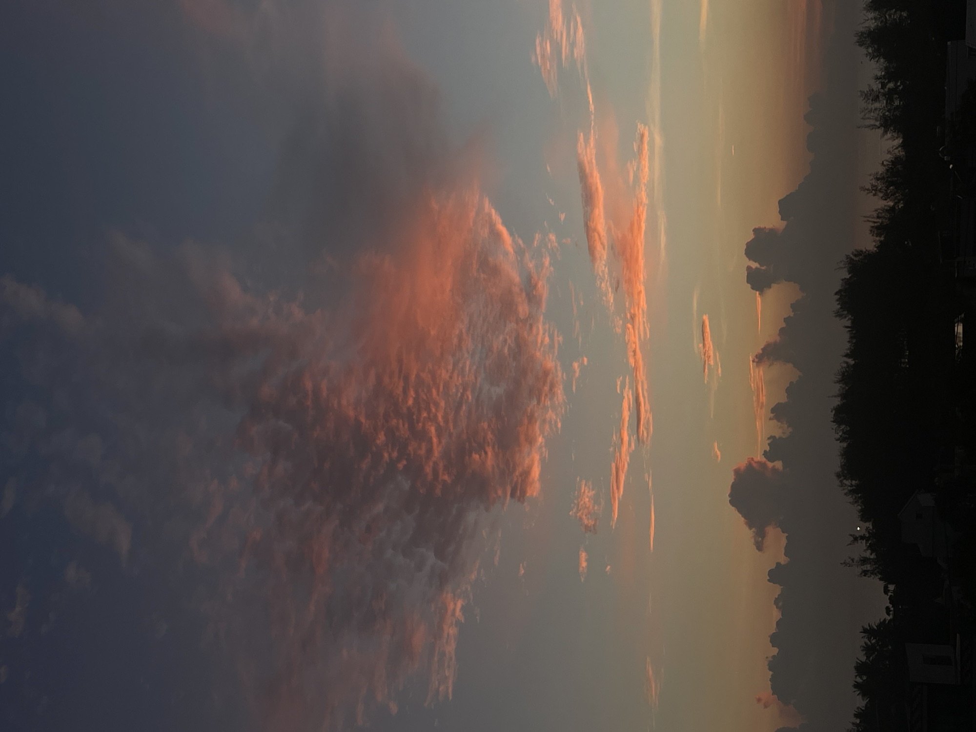 The image depicts a sunset sky. The left side of the image shows dark clouds, while the center and right sides feature clouds tinged with orange and pink hues, suggesting the sun's position below the horizon. In the far right, a silhouette of trees and buildings is visible against the twilight sky. There is no indication of the city of Đà Nẵng, Vietnam. There are no people, subjects, or actions visible in the image.