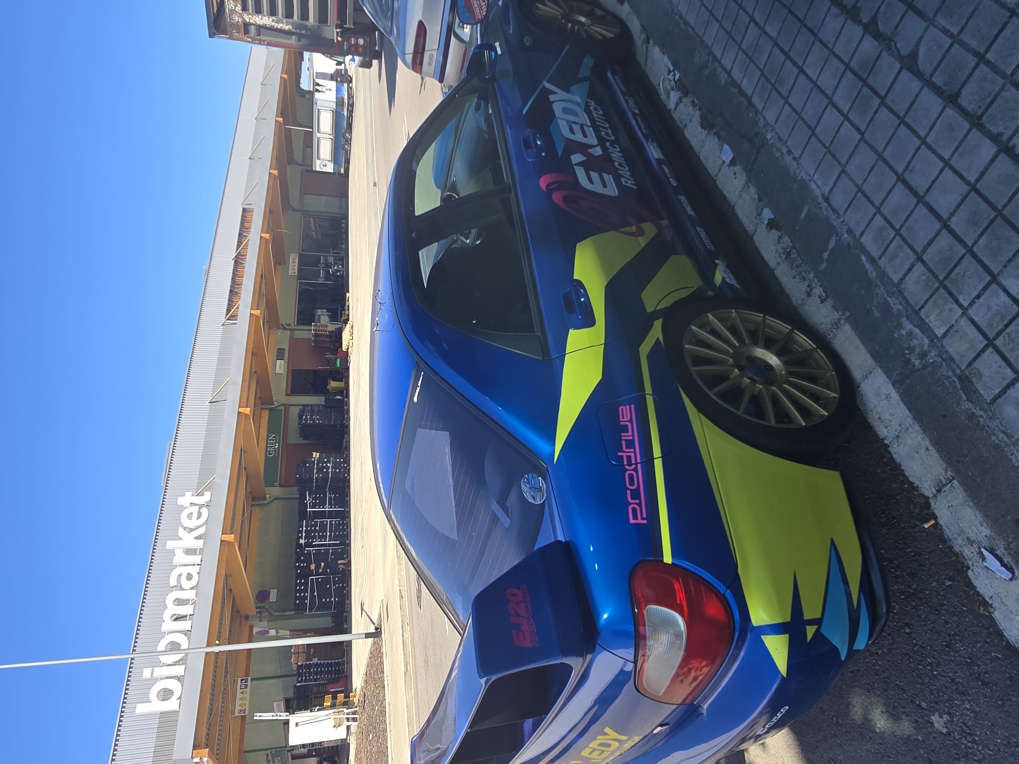 The image shows a blue Subaru Impreza WRX STI with racing decals parked on a street next to a Bioco Market in Barcelona, Spain. The car features sponsor logos, a prominent wing, and gold-colored wheels. The background includes the market's facade with the "Biomarket" sign visible, along with other commercial buildings and a clear blue sky. The street is paved with concrete tiles. There is a cigarette butt lying near the curb next to the car.