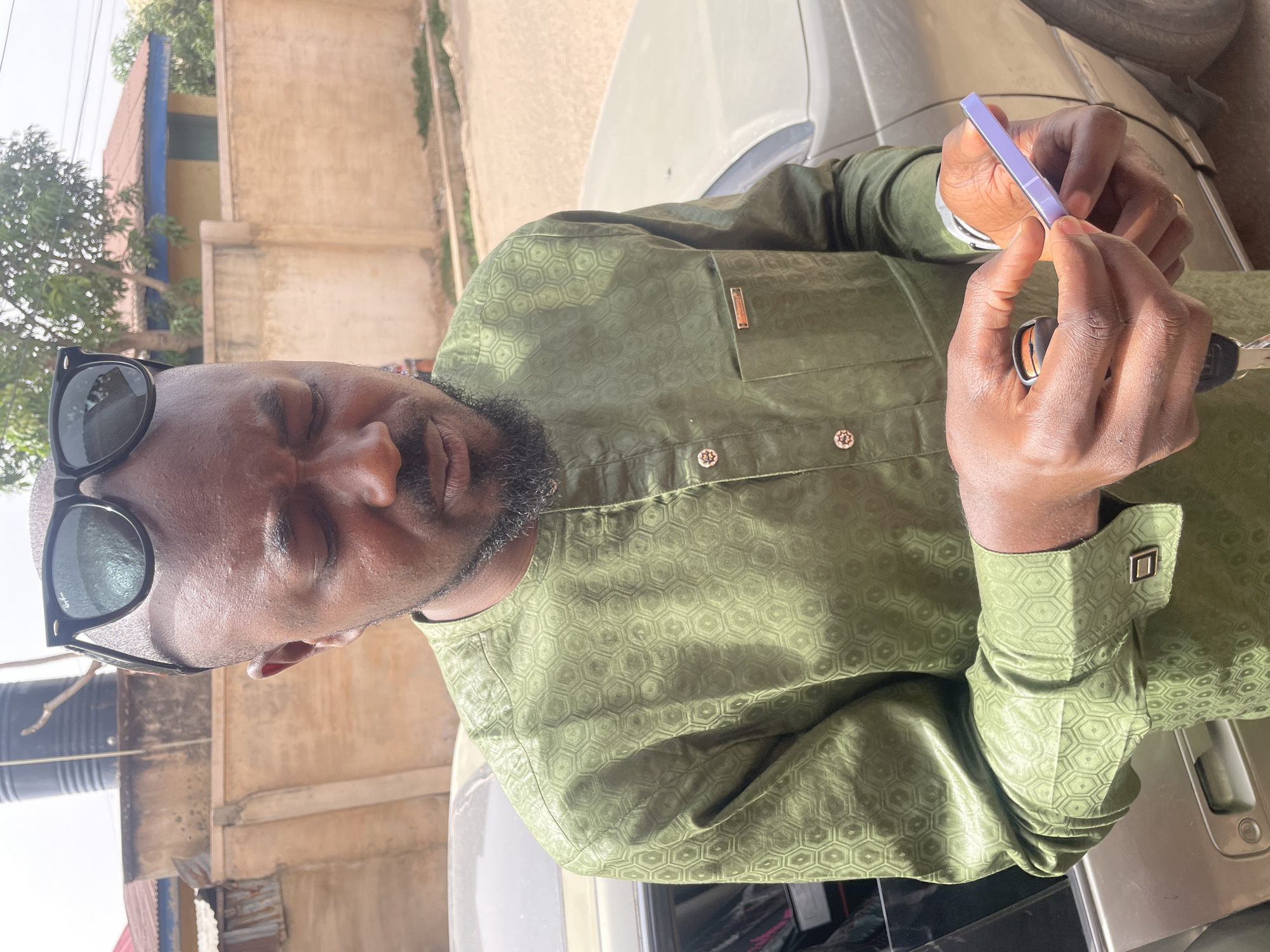A man with sunglasses on his head is holding a small, purple object and keys in his hands. He is wearing a patterned green shirt with cuff links. He appears to be standing near a vehicle. The background includes buildings and trees, suggesting an urban or suburban location. Based on the request, this location is presumed to be Jos, Nigeria.