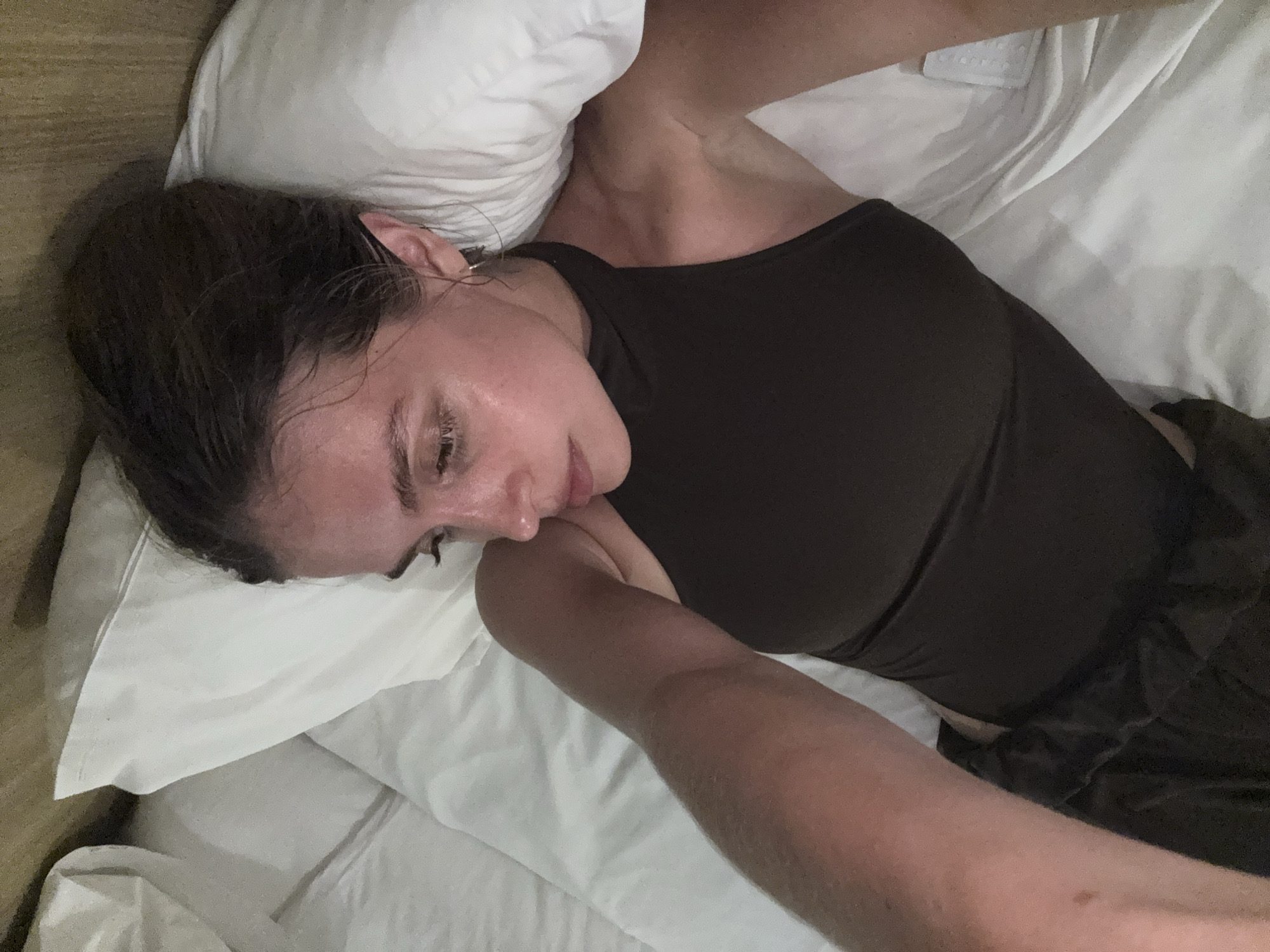 A woman lies in a bed in a room, possibly a hotel room. She is resting her head on a white pillow. She is wearing a dark-colored tank top. The environment appears to be a simple, indoor setting with basic bed linens. There is no direct indication of the location being Patong, Thailand, or any other specific geographic region within the image.