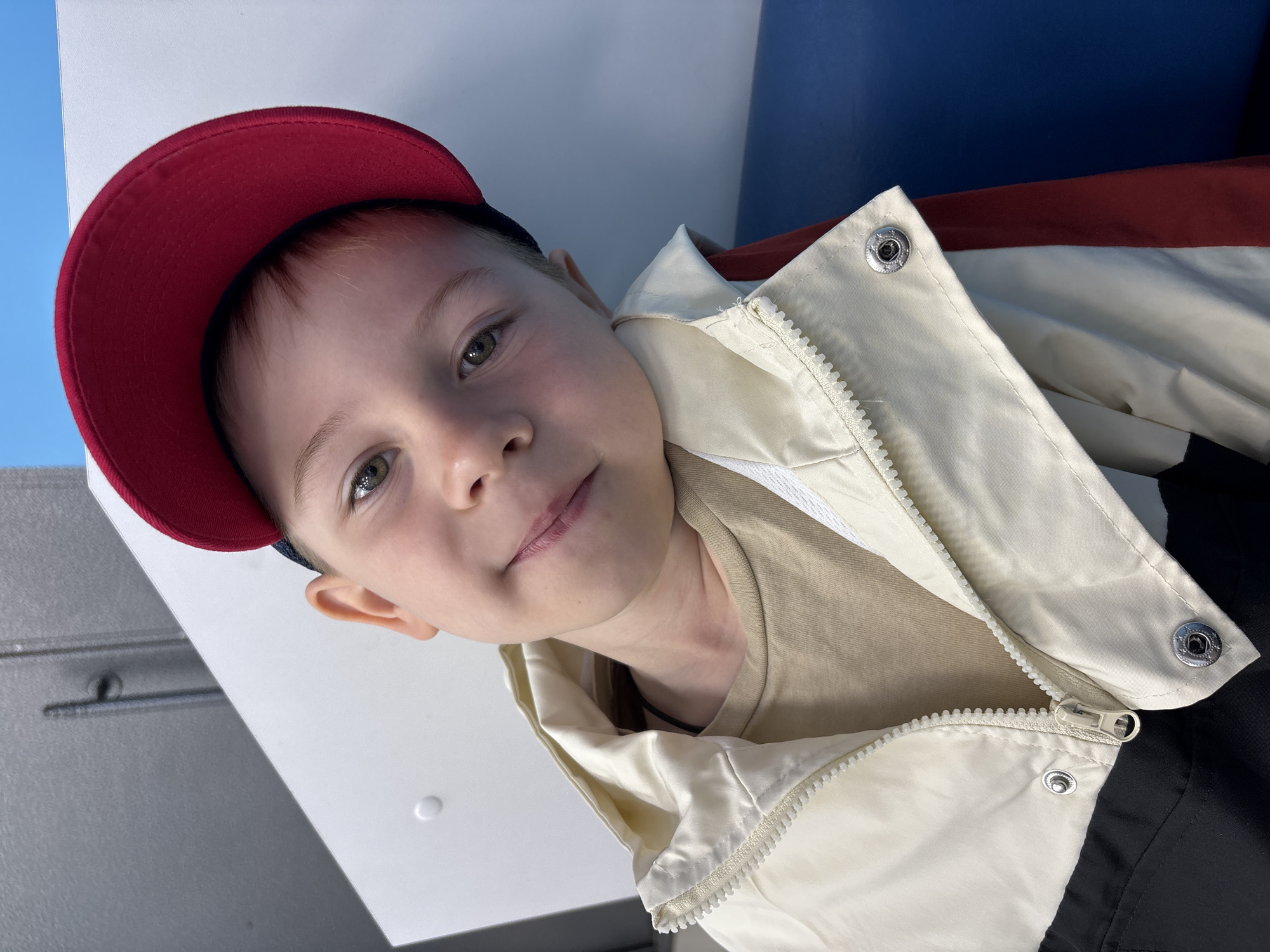 A young child with light skin, light brown hair, and green eyes is centered, looking directly at the camera with a slight smile. The child is wearing a red baseball cap, a light beige t-shirt, and a multi-paneled jacket that is cream/off-white on the upper portion, black on the lower portion, and includes a dark red stripe visible on the right side. The jacket features a white zipper and silver snap buttons. The child's head is tilted to the right. The background consists of a white vertical surface behind the child, a smooth blue surface to the child's right, and a light gray surface with a faint vertical line above and to the child's left. The scene is brightly and evenly lit. The location is Rusanovo, Russia.