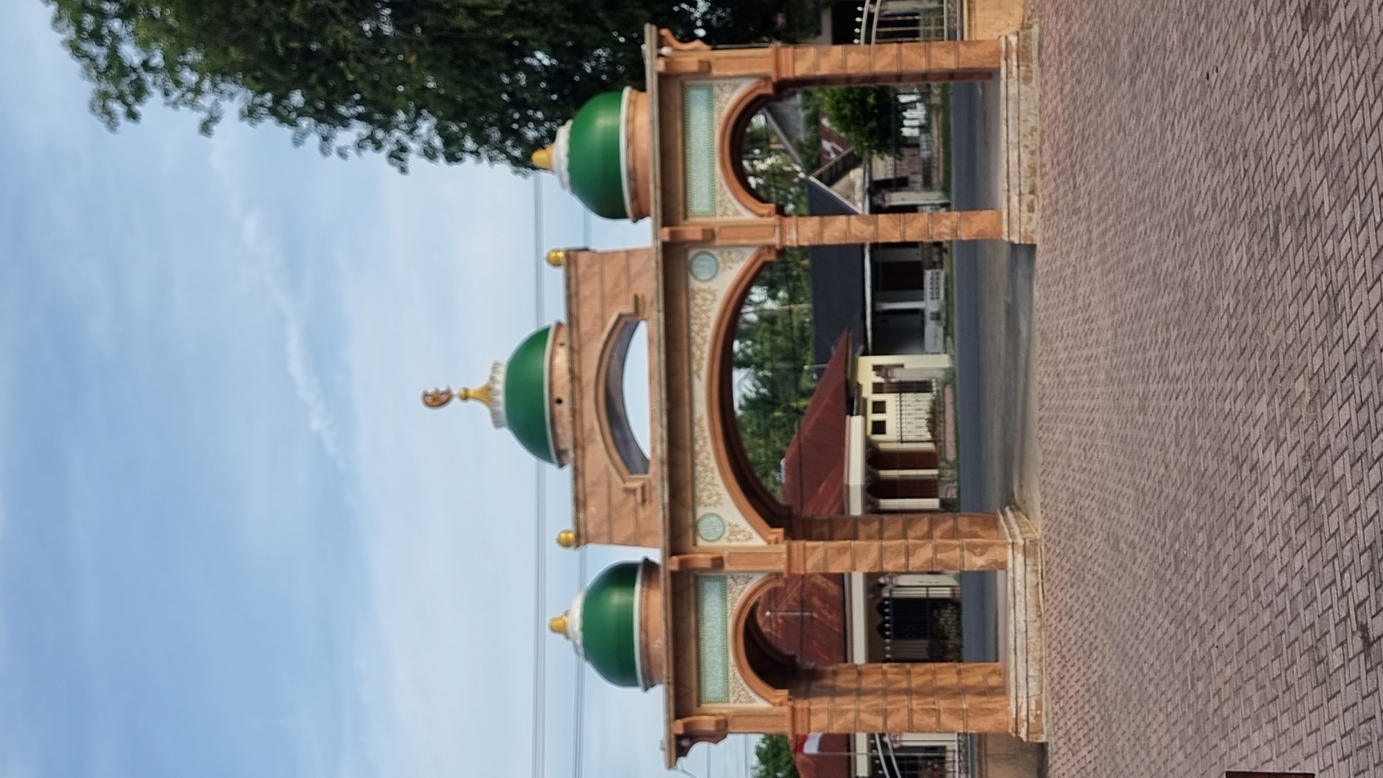The image depicts the entrance archway to a mosque in Meunasahlhok, Indonesia. The archway is constructed with a sandy-colored material, possibly brick or stone, and features two prominent green domes topped with gold crescent moons. The main archway is flanked by two smaller arched passageways, each also topped with a green dome and crescent moon. The ground in front of the archway is paved with reddish-brown brick pavers. In the background, through the arches, residential buildings with red and grey roofs are visible, along with some trees. The sky is cloudy and a light blue. There are no people visible in the image. The overall impression is of a religious and welcoming structure in a suburban or semi-rural setting.