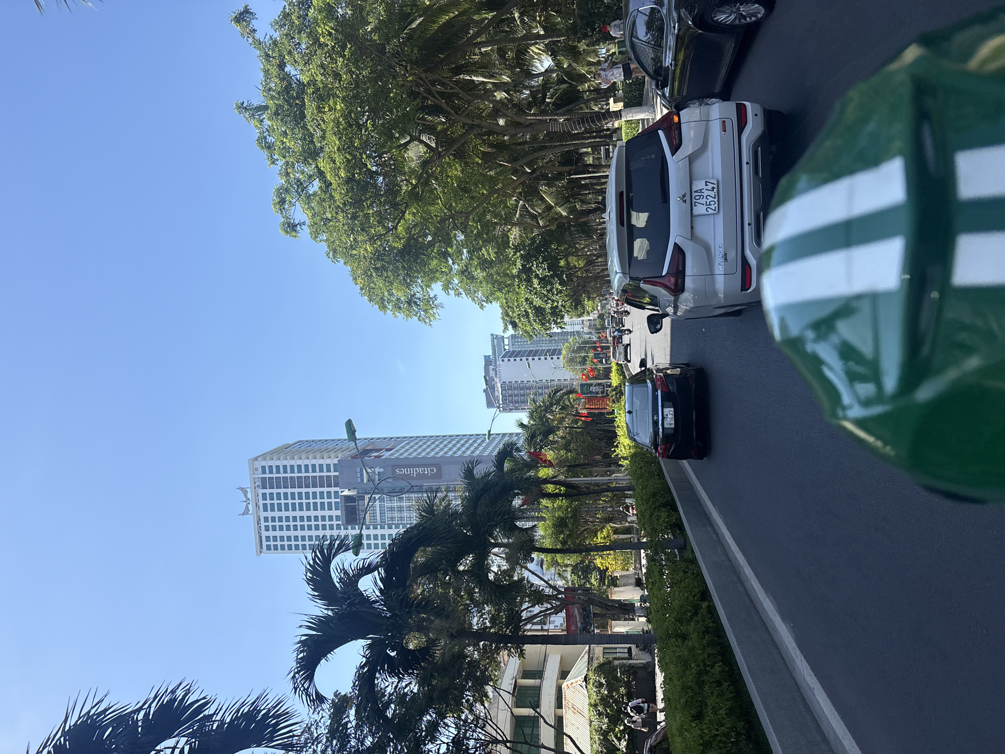 The image captures a street scene in Nha Trang Ward, Vietnam. A road runs along the right side of the frame, lined with a low hedge. A silver car is followed by a black car on the road, both facing away from the viewer. A green object with white stripes, likely part of a vehicle or object close to the camera, is prominent in the right foreground. Palm trees and other lush greenery line the road, with buildings visible behind the trees. In the distance, taller buildings, including one with "citadines" written on it, are visible against a clear blue sky. Other cars and possibly pedestrians are visible further down the street, creating a sense of urban activity.