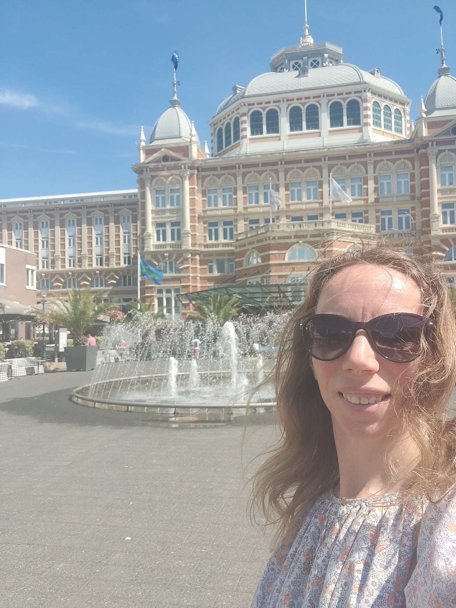 A woman with blonde hair, wearing sunglasses and a floral print dress, is taking a selfie in front of a large building with a domed roof and a fountain. The building, with its ornate architecture and red brick facade, is the Kurhaus Hotel, a historic landmark located on the Scheveningen beach in The Hague, Netherlands. The sky is clear and blue, indicating a sunny day. The fountain in the foreground is spraying water, adding a dynamic element to the scene. The woman appears to be smiling and enjoying her visit.