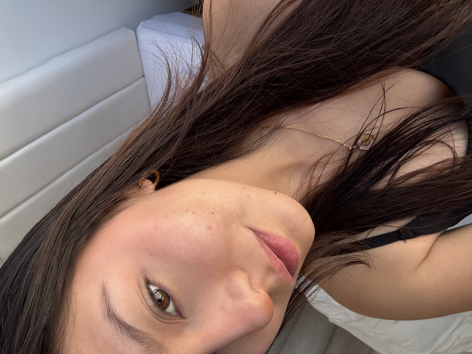 A close-up, angled portrait features a person, likely female, with long, dark brown hair and a light to medium skin tone. The subject's head is tilted, suggesting they are reclining or lying down, with their gaze directed towards the camera from a slightly elevated perspective.
The person's face is central, with one brown eye prominently visible, reflecting what appears to be an overhead light or window. Their skin shows natural pores and a few small moles or freckles. Their lips are softly parted, enhanced with a subtle pink hue. Long, dark brown hair frames the face and cascades around the shoulders.
A delicate golden chain necklace with a small, light-colored hexagonal pendant is worn around the neck. A black strap, likely from a top or bra, is visible on their left shoulder.
The background is simple, composed mainly of a white, cushioned surface featuring subtle horizontal seams, possibly a sofa, bed, or seating in a boat. A darker, indistinct area is visible in the upper right. The lighting is soft and natural, illuminating the subject's features. No text is visible. This scene is located in San Francisco Ocotlán (Ocotlán), Mexico.