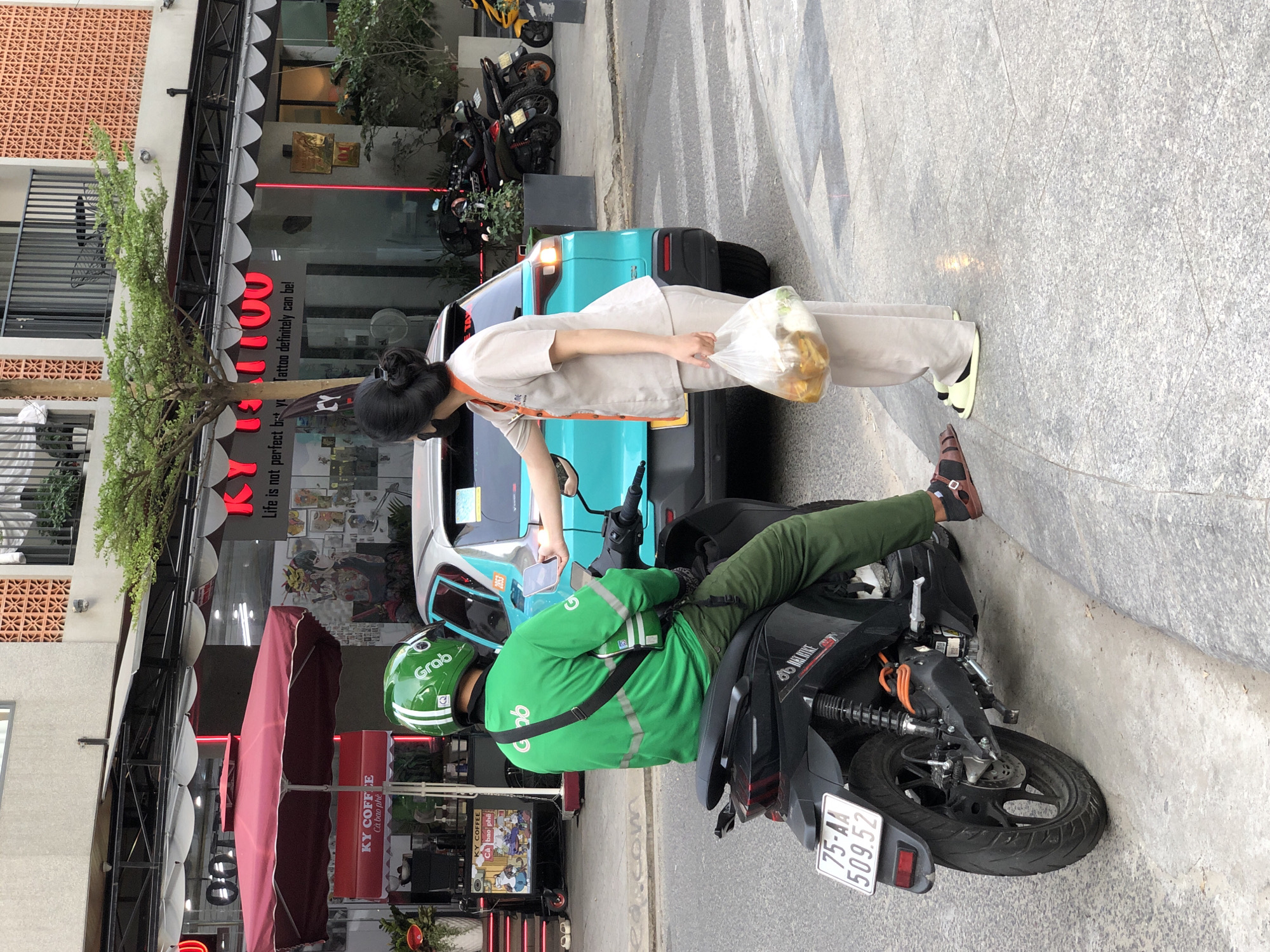 In the city of Đà Nẵng, Vietnam, on a paved street with a concrete curb, a male Grab delivery driver, wearing a green helmet with the Grab logo and a bright green long-sleeved shirt with reflective stripes, is seated on a black motorbike. The motorbike has a license plate reading "75-AA 509.52". The driver is interacting with a woman who is standing next to a turquoise car. The woman is wearing a light-colored uniform with orange trim and is holding a clear plastic bag filled with bread. She is handing a smartphone to the Grab driver, who is also holding a smartphone. In the background, there are buildings with shops, including a store with "KY COFFEE" signage and a tattoo parlor with neon red lettering. Several other motorbikes are parked further down the street.