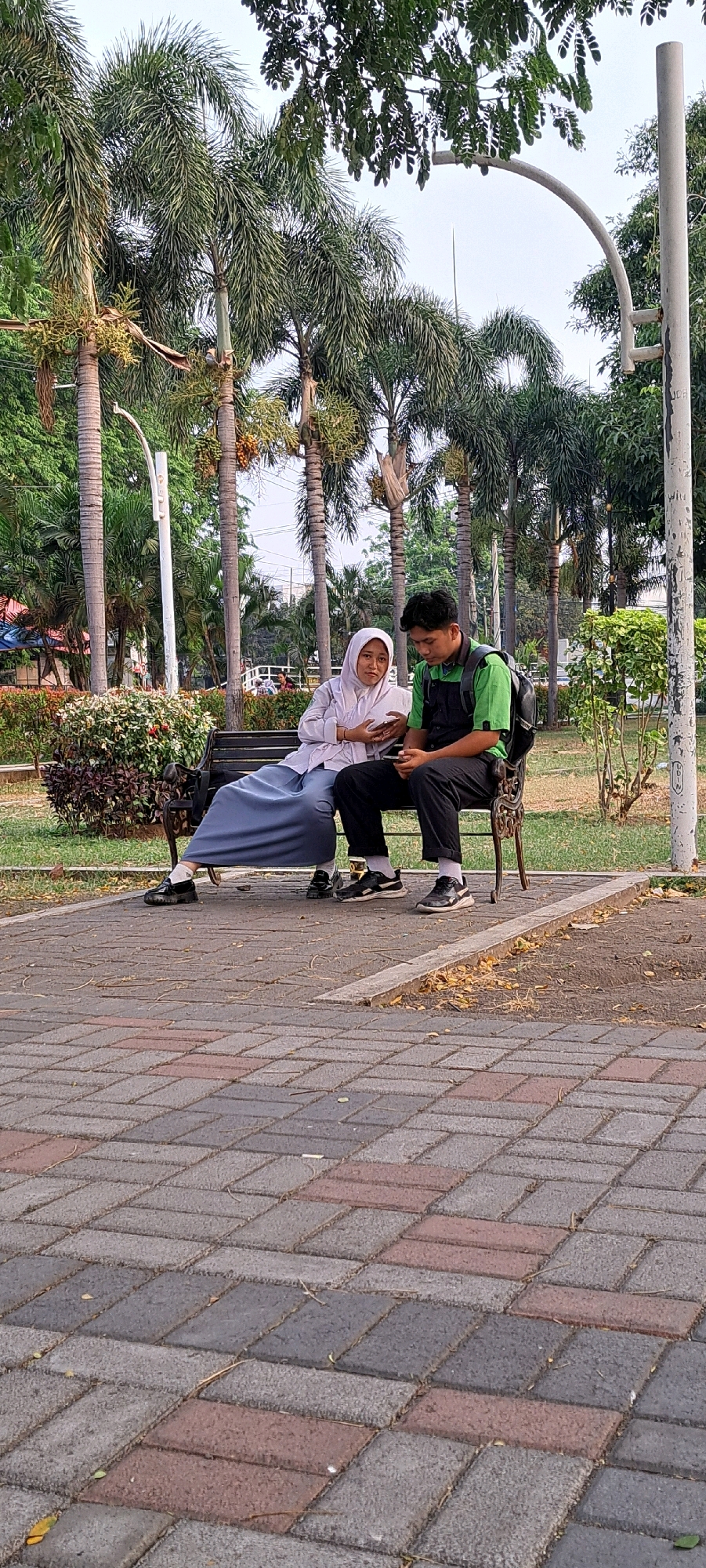 In the city of Bangil, Indonesia, two young people are sitting on a park bench under tall palm trees. The person on the left, wearing a hijab and a light-colored uniform, is looking at their phone. The person on the right, wearing a green shirt and black pants with a backpack, is also looking at their phone. They are sitting on a dark metal bench with ornate legs. The ground in front of the bench is paved with interlocking bricks in shades of gray and red. Lush green bushes are behind the bench to the left, and grass and more palm trees are visible in the background. A tall, curved white lamppost is to the right of the bench. The sky is overcast.