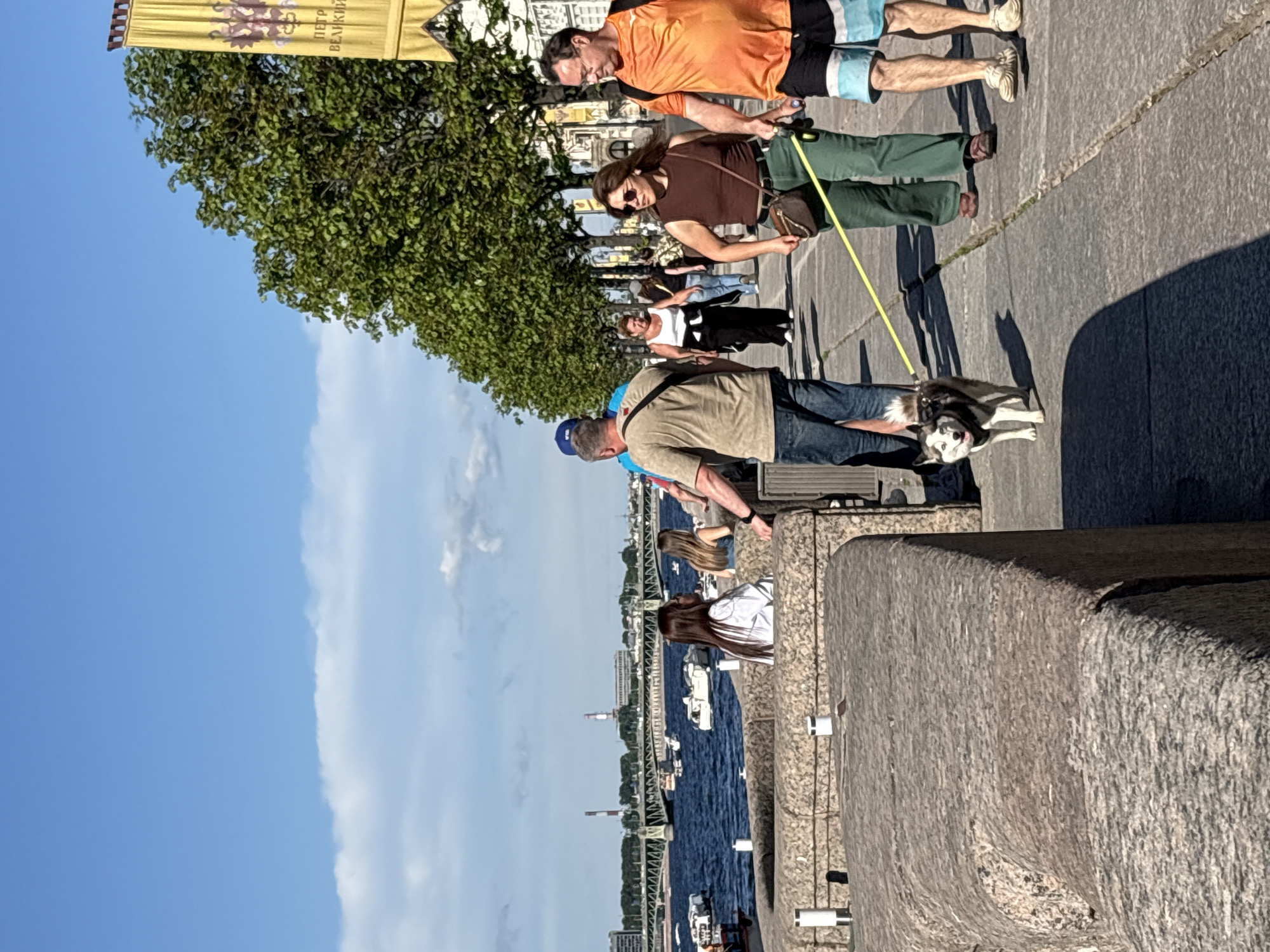 This image captures a vibrant sunny day scene on a granite embankment promenade in Saint Petersburg, Russia, likely along the Neva River, with the Liteyny Bridge visible in the distance.
In the foreground, a low, textured granite wall runs horizontally, with white-capped metal bollards integrated into it. A small husky dog, predominantly white with black and grey markings and blue eyes, walks on a yellow leash on the pavement directly in front of the wall, looking towards the viewer with its tongue slightly out. To the dog's left, a man in a beige short-sleeved shirt and blue jeans, with a dark bag across his chest, is partially visible, holding part of the leash. To the dog's right, a woman with dark hair wears a brown sleeveless top, green wide-leg pants, and sunglasses, carrying a brown shoulder bag and holding the yellow leash. To her right, a man in an orange t-shirt and blue-and-white patterned shorts walks with a slight forward lean.
Further along the promenade, a line of lush green trees casts shadows on the pavement. Several other individuals are walking in the midground; notable figures include a woman in a white tank top and black pants walking away from the viewer, and a man in a blue cap with his back to the viewer. On the far right, a tall, yellow banner with a double-headed eagle crest and the Cyrillic text "ПЕТР ВЕЛИКИЙ" (PETER THE GREAT) is partially visible.
In the background, the wide, dark-blue Neva River flows, dotted with numerous white recreational boats and smaller vessels. A large, green metal truss bridge spans the river, with visible arch structures and supports. Beyond the bridge, the cityscape includes various buildings and two prominent industrial chimneys on the far bank. The sky is clear and blue with scattered white, fluffy clouds. Two women are seated on the lower granite embankment wall facing the river, one with long dark hair wearing a white long-sleeved shirt, observing the scene.