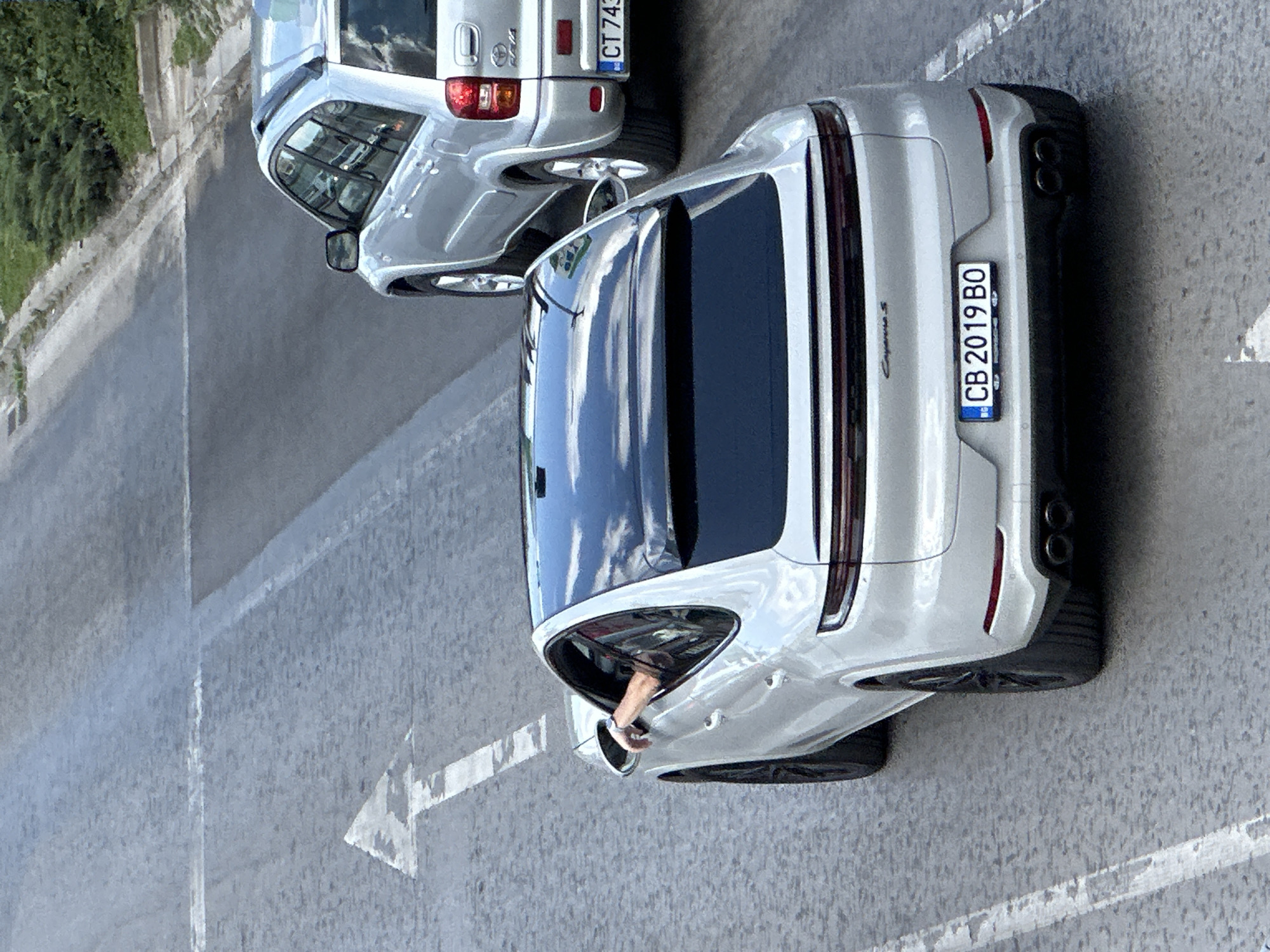 The image shows two vehicles on a street in Veliko Tarnovo, Bulgaria. The foreground is dominated by a silver Porsche Cayenne Coupe, its rear facing the camera. The car has a black panoramic roof and dual exhaust pipes. Its license plate reads "CB 2019 BO". A person's arm is visible resting on the open driver's side window. Behind the Porsche, a silver SUV, possibly a Toyota RAV4, is also present, with its license plate reading "CT 743". The vehicles are on a paved road with white lane markings. In the background, a sidewalk and green foliage are visible. The overall scene suggests a normal urban traffic situation.