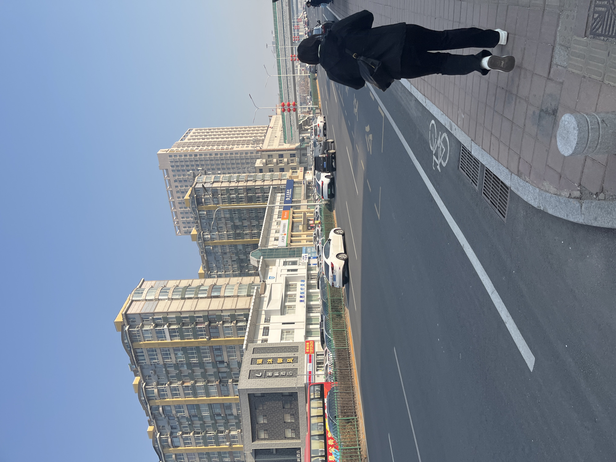 An outdoor urban scene in Changchun City, China, depicts a person walking along a sidewalk parallel to a busy street lined with multi-story buildings under a clear blue sky. The image, oriented correctly, shows the person in a black jacket and pants with white sneakers, walking away from the viewer on the right side of the frame. To their left is a paved road with multiple lanes, including a bicycle lane marked with a bicycle symbol. Several cars, including a white sedan, a black SUV, and other vehicles, are visible on the road, suggesting moderate traffic. In the background, an elevated overpass or bridge can be seen, with red lanterns decorating some of the street poles.
The left side of the image is dominated by tall, modern residential and commercial buildings. Some of the buildings feature numerous windows and air conditioning units. At street level, various businesses are housed in these buildings. Visible text includes signs for "丽枫酒店" (Lavande Hotel), "枫酒店" (Maple Hotel), "瑶沐御足" (Royal Foot Bath/Spa), "恩黑医疗美容" (Enhei Medical Aesthetics), and "吉林农商银行" (Jilin Rural Commercial Bank), along with a "旺铺出租" (Shop for Rent) sign and a phone number (13019128429). A red and white bus is also parked near the sidewalk. The time of day appears to be daytime with bright, clear weather.