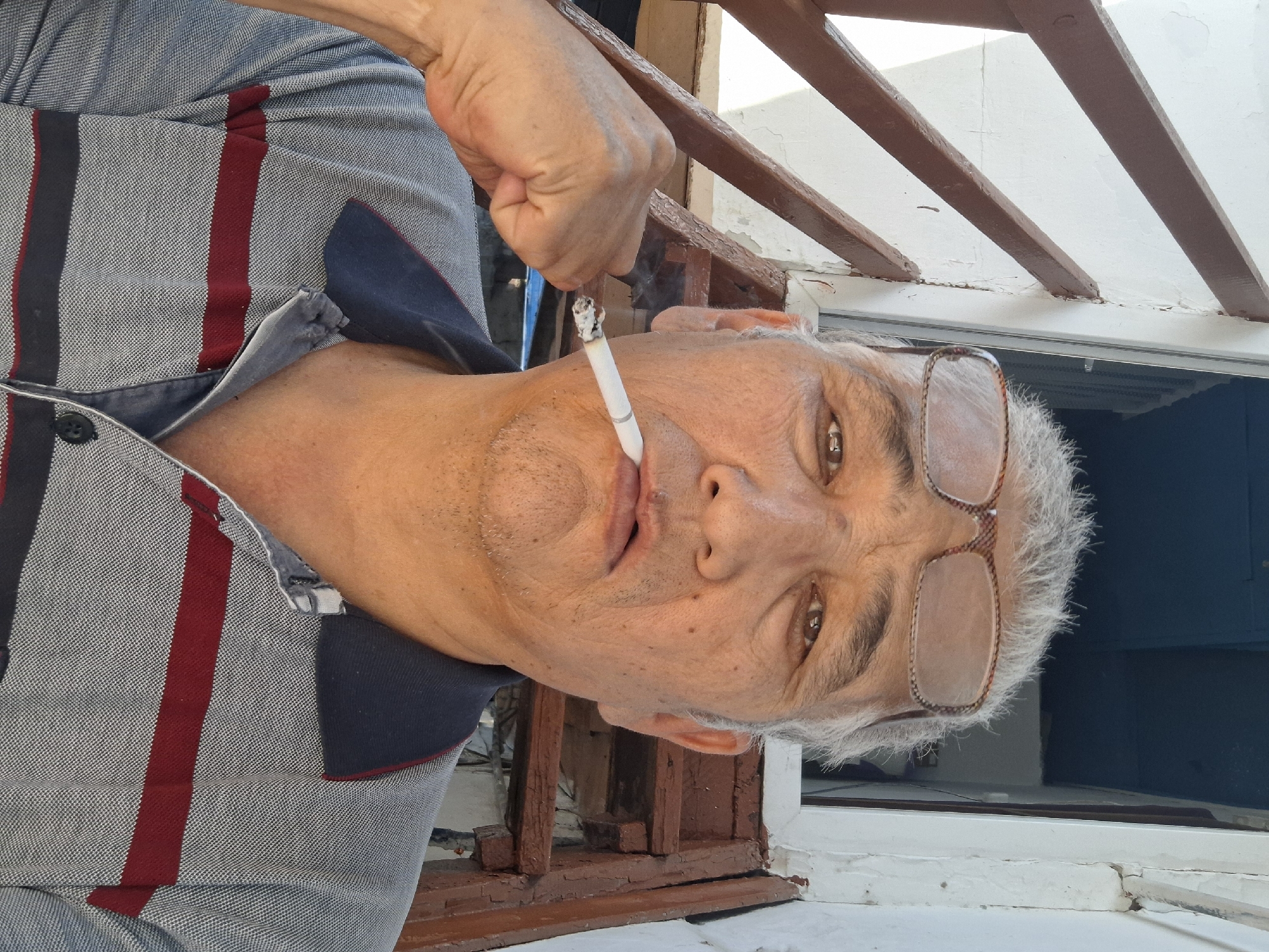 A middle-aged man with graying hair and glasses is smoking a cigarette. He is wearing a striped polo shirt and appears to be outdoors, possibly on a balcony or near a staircase. The background suggests an urban setting, possibly a residential area in Almaty, Kazakhstan, with some visible architectural elements.