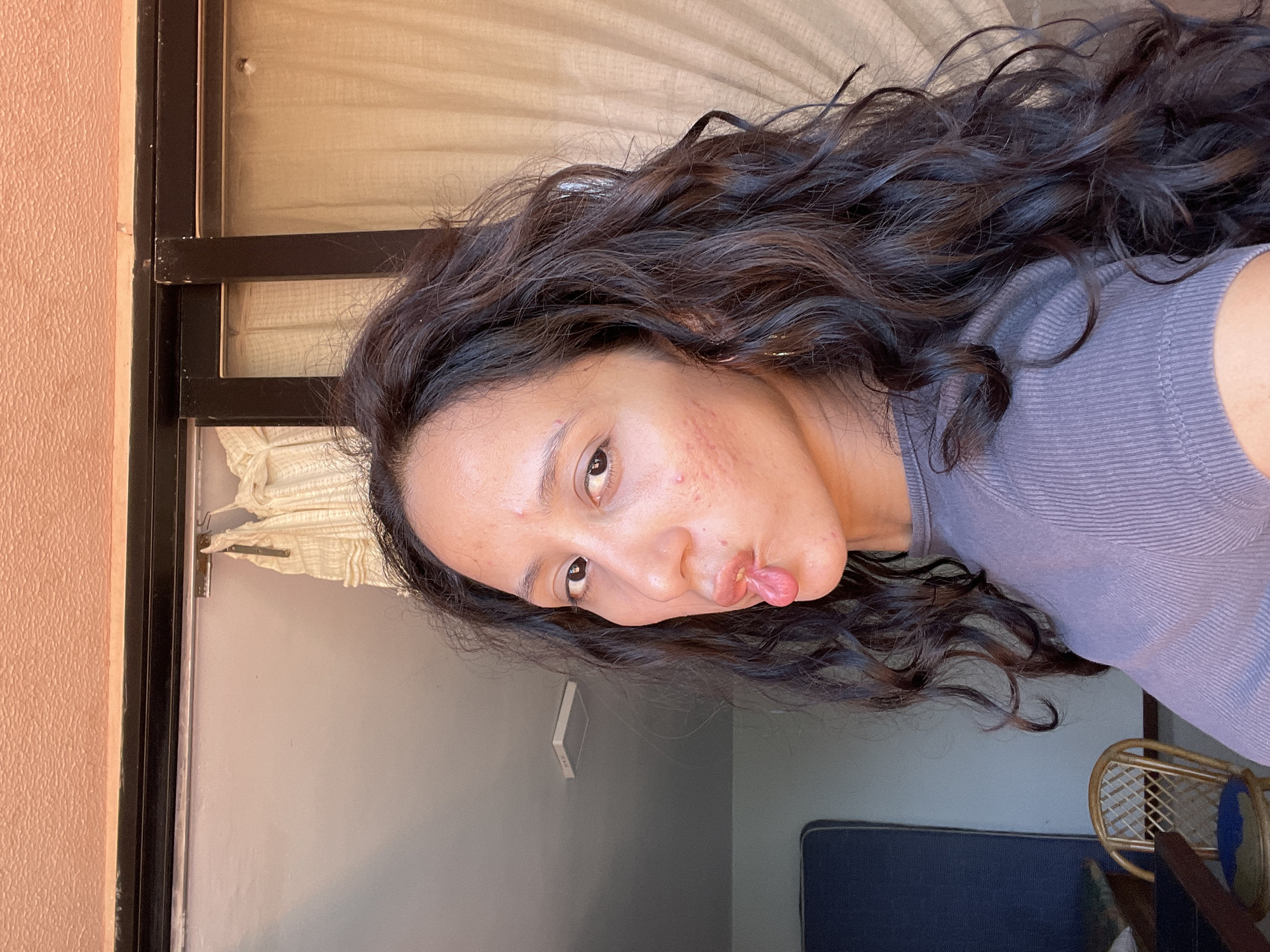 A woman with dark, wavy hair, wearing a purple t-shirt, is taking a selfie. She is sticking her tongue out to the side, with her lips pursed. Her face is turned to the right, and her eyes are looking towards the camera. She has some acne on her cheeks. The background appears to be an indoor setting with a window that has sheer curtains. Behind her, to the left, is a blue mattress and a wicker chair. The ceiling is textured and orange. The scene is brightly lit, suggesting natural light. The image is a close-up of the woman, with the background slightly out of focus. The location is identified as Porlamar, Venezuela.