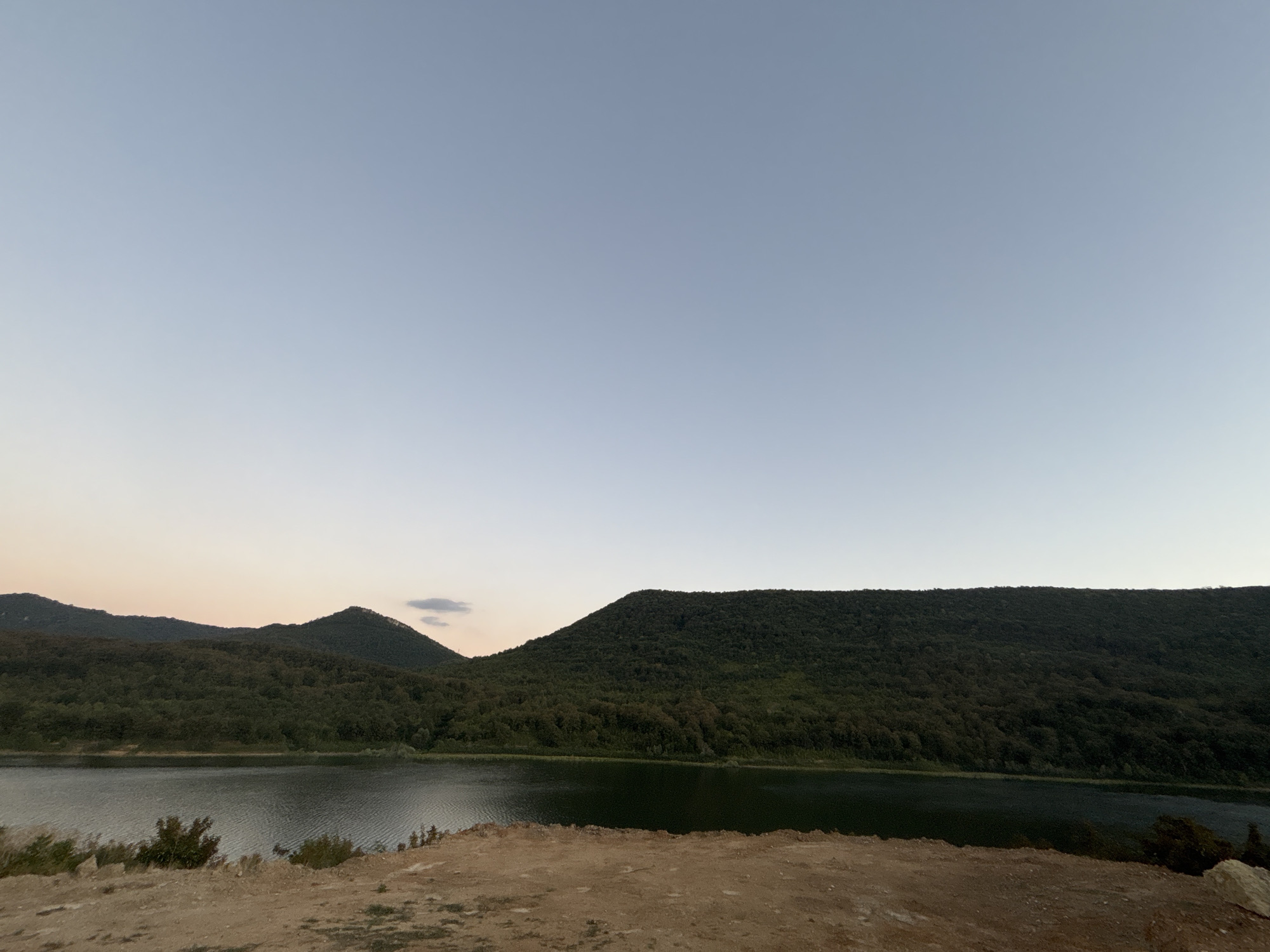 This image depicts a serene landscape with a body of water in the foreground, bordered by lush green hills and mountains in the background. The sky is a pale, clear blue, suggesting either early morning or late evening. The terrain in the immediate foreground appears to be dry, sandy earth with sparse vegetation. The water has a slight ripple, reflecting the muted light. The hills are densely covered with trees, creating a dark green, textured surface. The overall impression is one of natural tranquility. This scene is located in the city of Asparuhovo, Bulgaria.