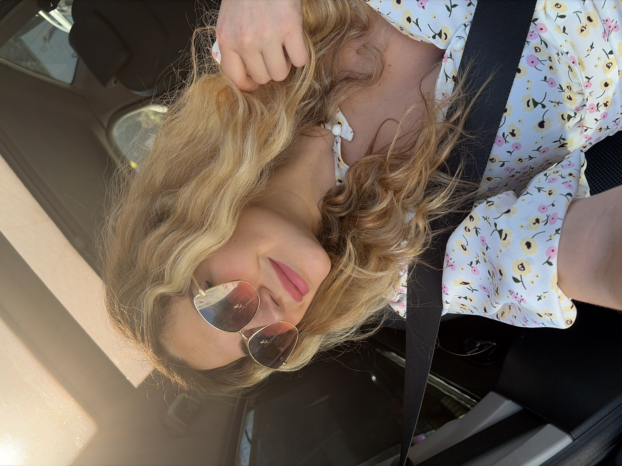 A blonde woman with wavy hair is sitting in a car, wearing sunglasses and a floral dress. The sun is shining through the sunroof, casting a warm glow on her face. She is smiling and touching her hair. The seatbelt is visible across her chest. The car's interior is dark, with some reflections visible on the windows. The scene appears to be set in Budva, Montenegro, possibly during a sunny day.