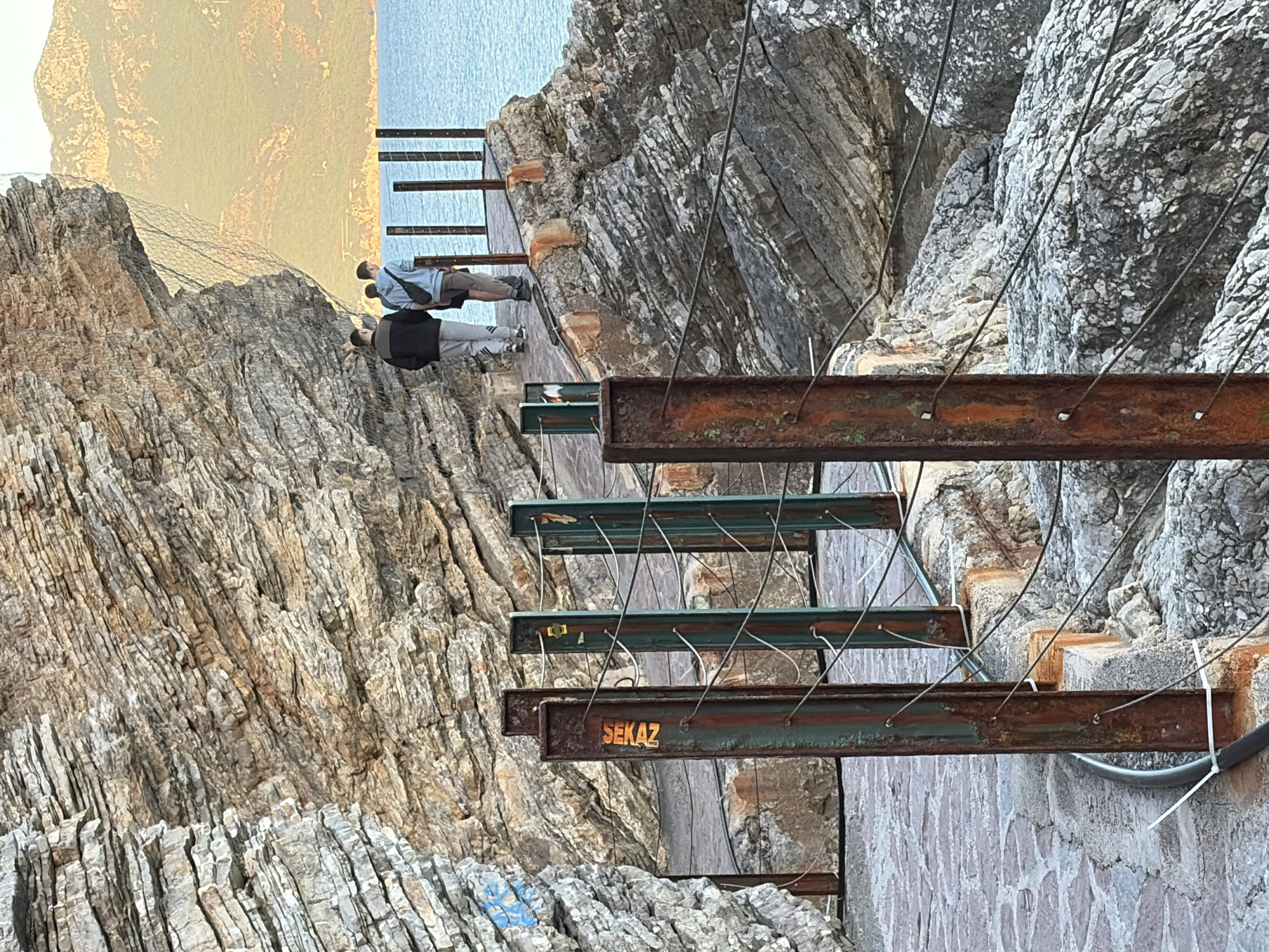 This media file captures a precarious-looking walkway built into a rugged cliff face in Budva, Montenegro, likely during the golden hour of early morning or late afternoon. The image is taken from a slightly elevated perspective, looking along the path.
The main subjects are two male individuals traversing the metal walkway. The person closer to the viewer is wearing a dark jacket and light grey pants, while the person slightly ahead is in a light blue shirt, dark shorts, and carrying a light shoulder bag. They appear to be walking or standing, observing the scenic coastal environment.
The scene features a steep, layered rock cliff face that dominates the left side of the frame. In the background, across the calm, blue sea, distant land or mountains are bathed in a warm, golden light. A wire mesh netting for rockfall protection is visible draped over the upper part of the cliff.
The most prominent objects are the metal beams forming the walkway, anchored directly into the rock. These structures show signs of age and exposure, with some sections appearing rusty brown (like I-beams) and others painted a dark green or teal. Thin metal wires are strung horizontally between these beams, serving as rudimentary safety barriers or guardrails. Some of these wires are secured with white zip ties to a lower beam. At the bottom of the image, a constructed stone wall forms part of the cliff's base.
A notable detail is the word "SEKAZ" painted in orange on one of the rusty metal beams, accompanied by a small, indistinct logo. There is also some faded blue graffiti on the lower rock face. The cliff itself exhibits intricate geological layering and texture.