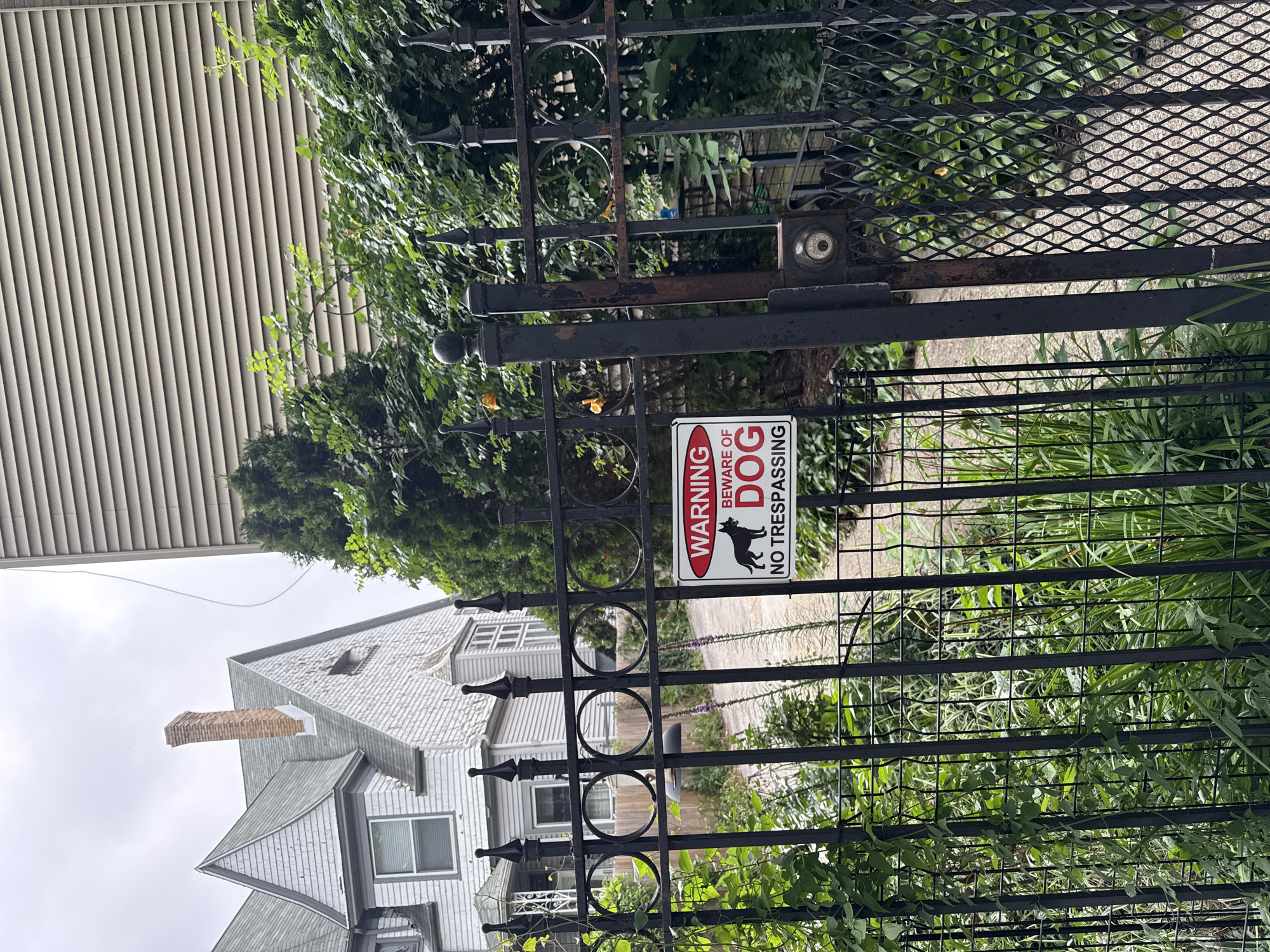 An exterior shot captures a black metal fence in front of a house. The fence has an ornate design with pointed finials and circular accents. A "WARNING BEWARE OF DOG NO TRESPASSING" sign is attached to the fence. Beyond the fence, a walkway leads to a light-colored, multi-story house with a grey roof and a brick chimney. Green foliage and trees are visible throughout the scene. The sky is overcast. This scene is located in Chicago, United States of America.