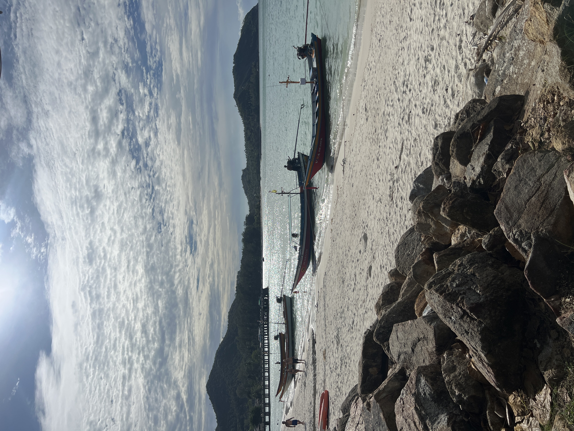 A serene, sun-drenched beach scene unfolds under a partially cloudy sky. The foreground is dominated by a cluster of large, weathered rocks, their rough textures contrasting with the fine, light-colored sand stretching towards the turquoise water. Several longtail boats, traditional Thai vessels, are moored near the shore, their colorful hulls reflecting the bright sunlight. In the mid-ground, a few individuals are visible on the beach, some walking along the water's edge, while others are near or in the boats. A pier extends into the distance from the left side of the frame, and beyond that, a lush, green island rises from the sea, its silhouette softened by the hazy light. The water is calm, with gentle waves lapping at the shore, and the sunlight creates a shimmering path across its surface.