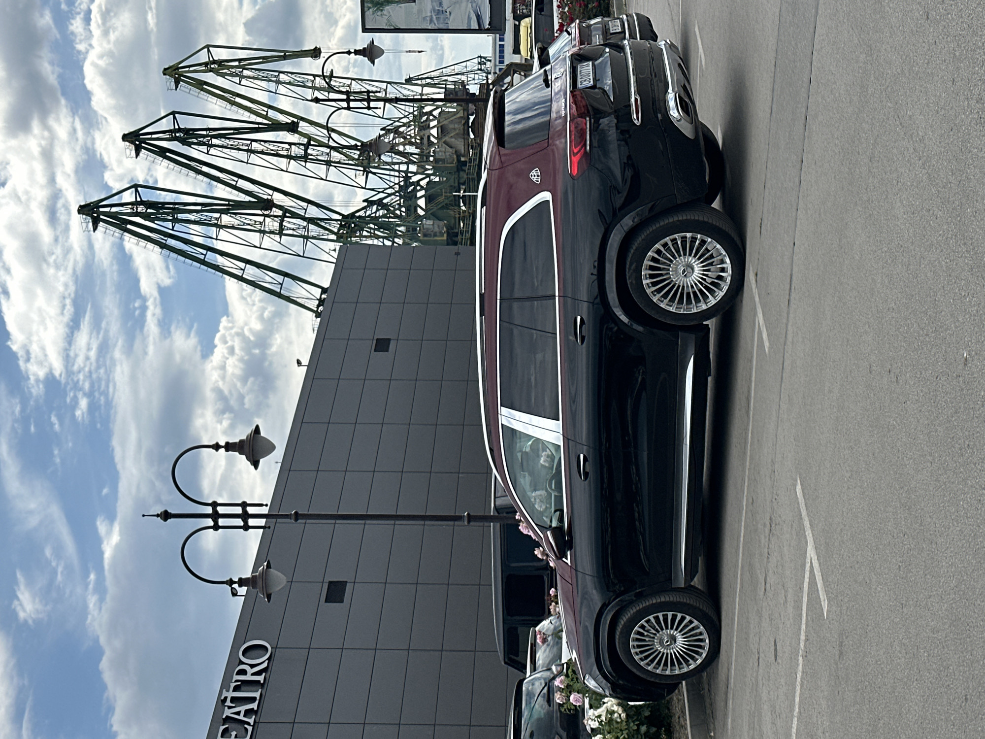 The image shows a black and maroon Maybach parked on the right side of the frame, partially obscuring the view. In the background, there is a modern gray building with the word "TEATRO" vertically written on the wall. Beyond the building, several green harbor cranes are visible against a sky filled with white clouds. Street lamps stand in front of the building. This photo was likely taken near the Varna Opera House in Varna, Bulgaria, due to the presence of the Teatro sign and harbor cranes in the background, common features of the area.