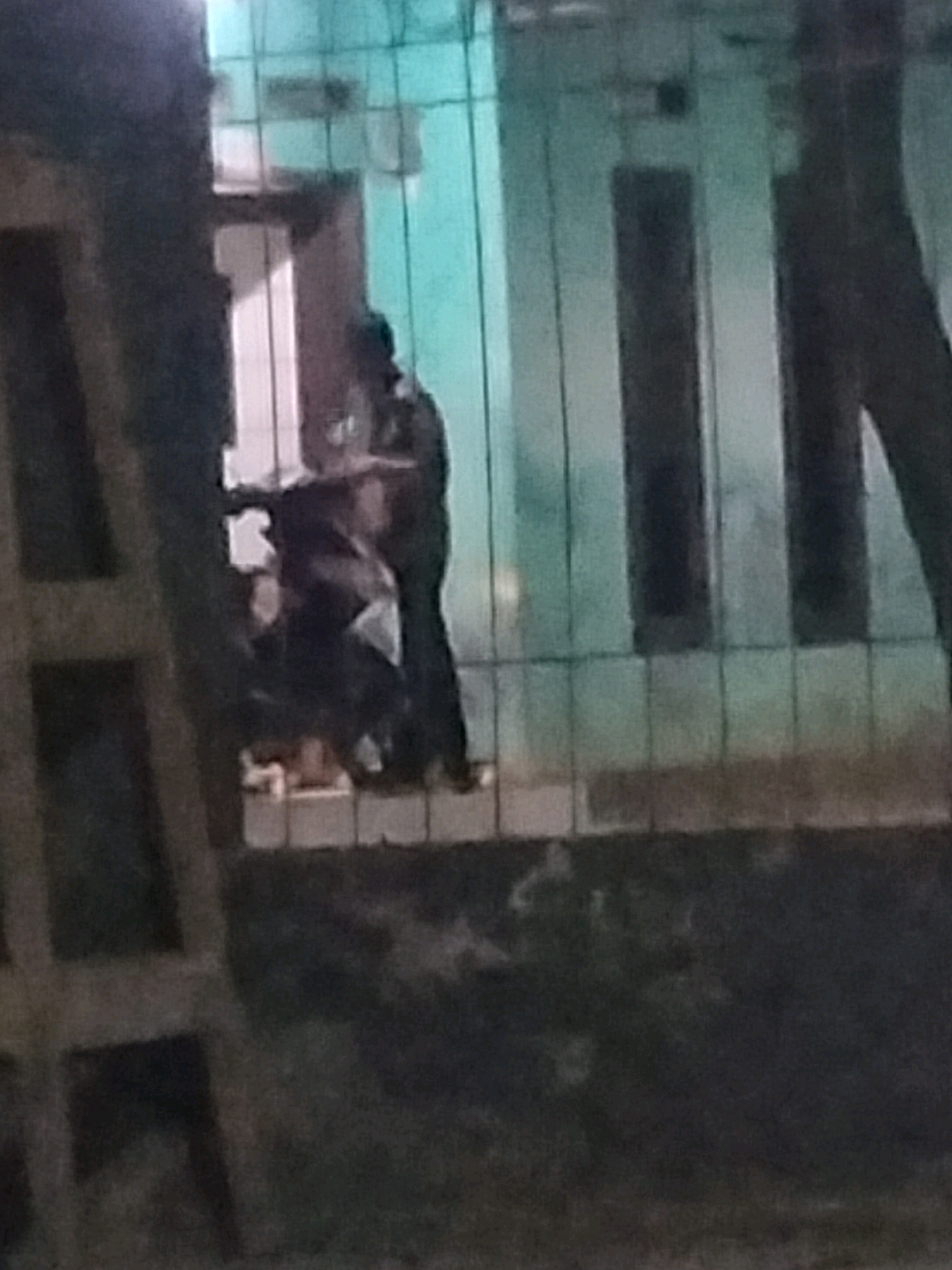 The image shows a dimly lit outdoor scene at night, possibly in Suka Sari, Indonesia. In the foreground, a partial view of a wooden structure, possibly a ladder or scaffolding, is visible on the left. Behind this, a building with a light blue wall and vertical dark bars across its facade is present. Two figures are discernible in the mid-ground, standing near the entrance of the building. One figure, taller and appearing to be wearing darker clothing, is standing upright. The second figure is partially obscured and appears to be interacting with the first. The ground in the foreground appears to be dark and uneven, with some patches of lighter color, possibly grass or debris. The overall lighting is low, creating shadows and obscuring fine details.