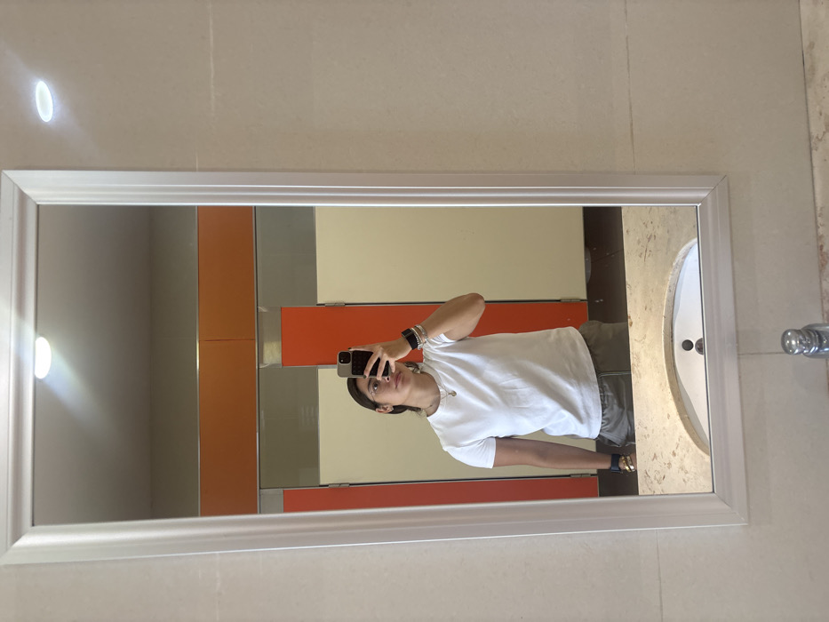 A person stands in front of a mirror in a tiled bathroom, taking a selfie with a smartphone. The reflection reveals a white t-shirt, olive-colored pants, and dark sandals. Behind the person, there are a set of lockers colored white, orange, and gray. A sink and faucet are partially visible to the right of the individual. The mirror has a white frame, and a light fixture is located near the top.
