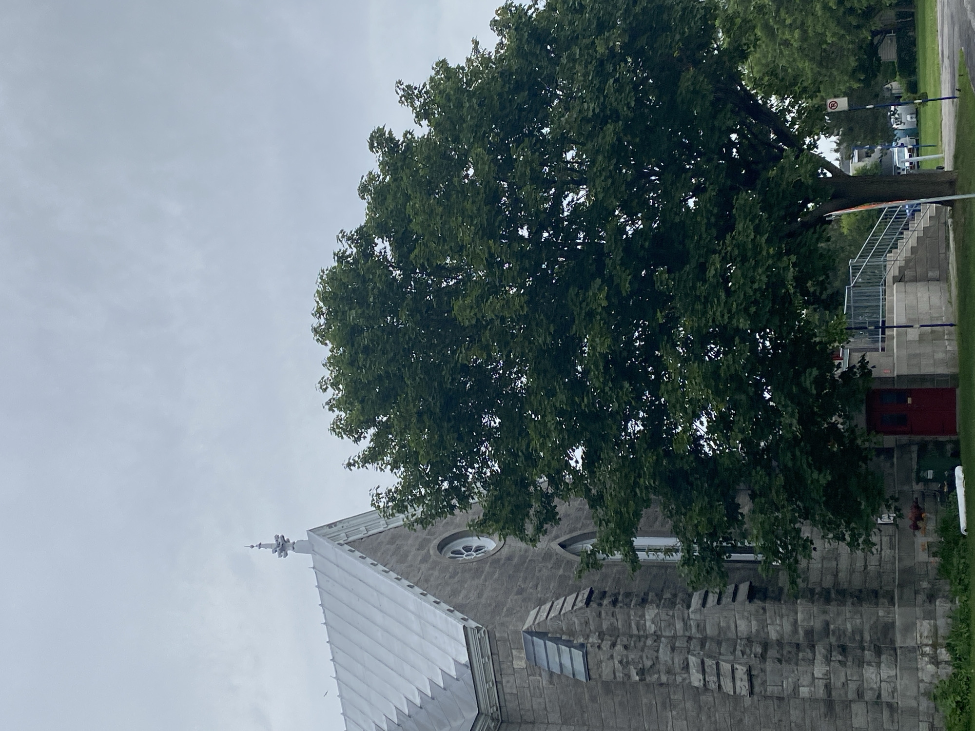 In Pointe-Claire, Canada, a stone church with a silver, stepped roof is partially obscured by a large, leafy green tree in the foreground. The church has a pointed spire topped with a decorative finial and features circular and arched windows. A red double door is visible on the side of the building. Adjacent to the church, a concrete staircase with metal railings leads up to an entrance. The sky is overcast. A green garbage bin and some scattered debris are present near the base of the church. Further in the background, other trees, a street, vehicles, and a residential building are visible. A "No Parking" sign with a circle and a diagonal line is posted on a pole.