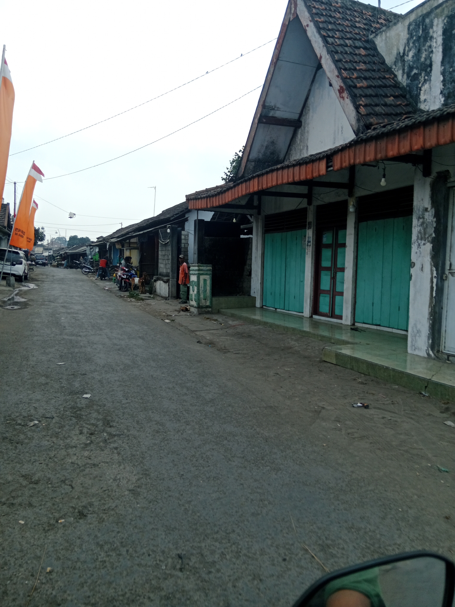 The image depicts a street scene in Sambi, Indonesia, with a row of shops lining the right side of the road. The shops appear to be closed, with green wooden shutters covering their entrances. The buildings are made of concrete and have tiled roofs. A person in an orange shirt stands on the sidewalk in front of one of the shops, next to a green pillar. A dog lies on the ground near the person.
On the left side of the street, there are several parked motorcycles and a white car. Flags, likely Indonesian flags with orange backgrounds and red and white elements, are planted along the side of the road. The road itself is paved and appears somewhat worn. The sky is overcast. The overall impression is of a quiet street, possibly in the early morning or late afternoon, with businesses closed.