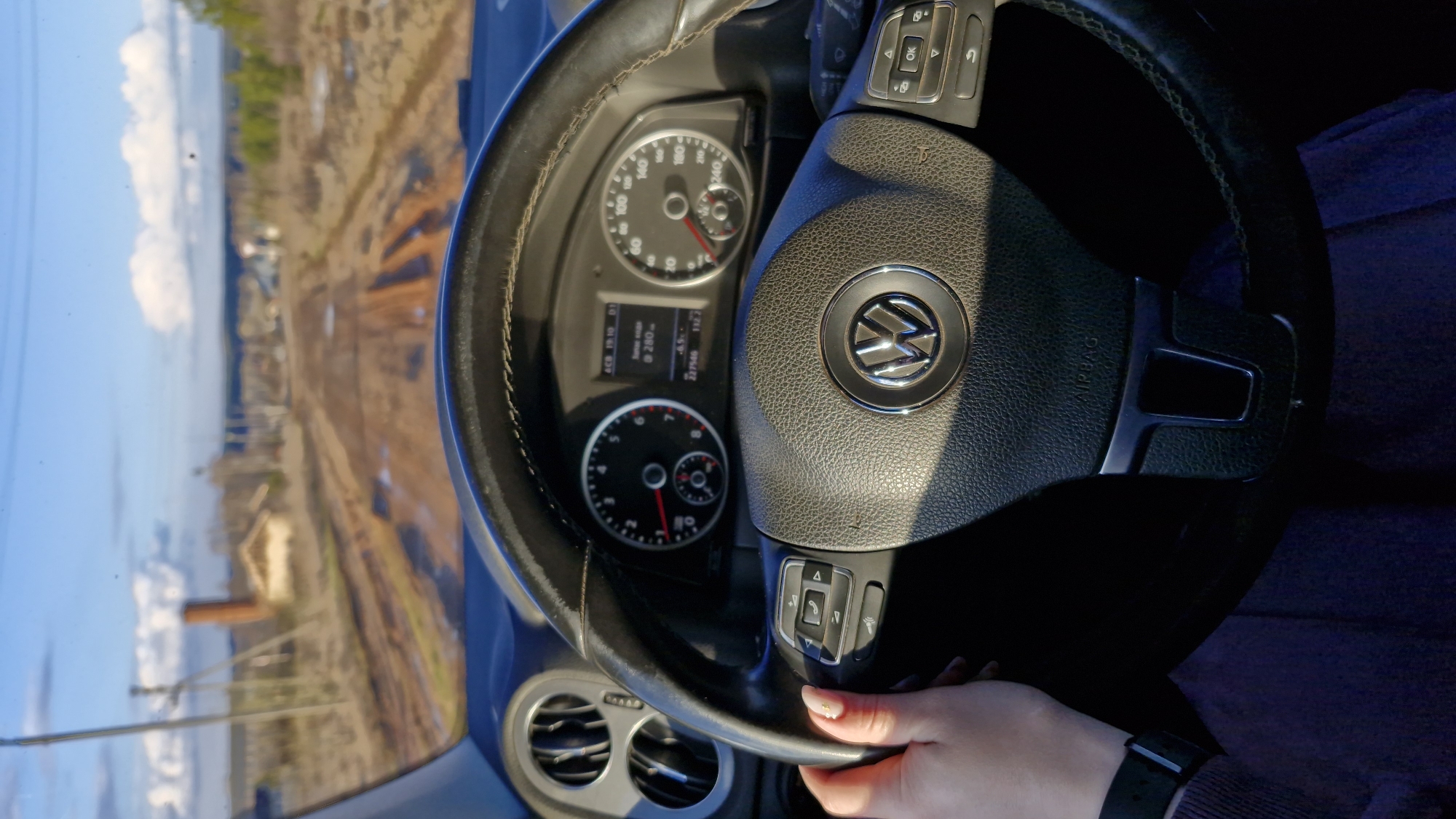 The image presents a first-person perspective from inside a vehicle, likely in Otchor, Russia, looking out through the windshield during daylight hours. A person's right hand, adorned with light-colored fingernails and a black smart-watch, rests on the lower right side of a black leather-wrapped Volkswagen steering wheel.
The car's dashboard is visible behind the steering wheel, featuring an analog speedometer, which reads approximately 60 units (likely km/h or mph), and a tachometer. A digital display screen shows text including "ACB 19:10 D1", "@280-", "-6.5C" (suggesting an outside temperature of minus 6.5 degrees Celsius), "227546" (possibly the odometer reading), and "1122". The steering wheel prominently displays the Volkswagen logo and features control buttons, including those for a phone and navigation, with "OK" visible on one button. The word "AIRBAG" is embossed on the steering wheel's hub. Air vents are visible on the dashboard to the left.
Outside the car, a bright, sunny sky with scattered white clouds dominates the upper half of the view. The landscape below is mostly an earthy, light brown terrain, resembling a dirt road, unpaved track, or industrial ground stretching into the distance. Distant structures, including a building with a prominent reddish-brown chimney or stack, and sparse, bare trees are visible, suggesting a potentially industrial or rural setting.