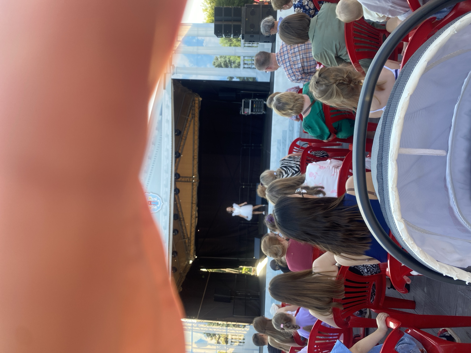 The image depicts an outdoor performance or event taking place on a stage, viewed from the perspective of an audience member. A young girl in a white dress is standing on the stage, appearing to perform. The stage has a black backdrop and is set against a white, classical-style structure with columns. In front of the stage, an audience is seated on red chairs. The audience members are diverse in age and gender, with many facing the stage. The foreground of the image is partially obscured by a blurry, pinkish-orange object, possibly a hand or finger. The overall setting suggests a public event in a park or open-air venue, possibly during daytime given the lighting. The location of Minsk, Belarus is not discernible from the visual information in the image.