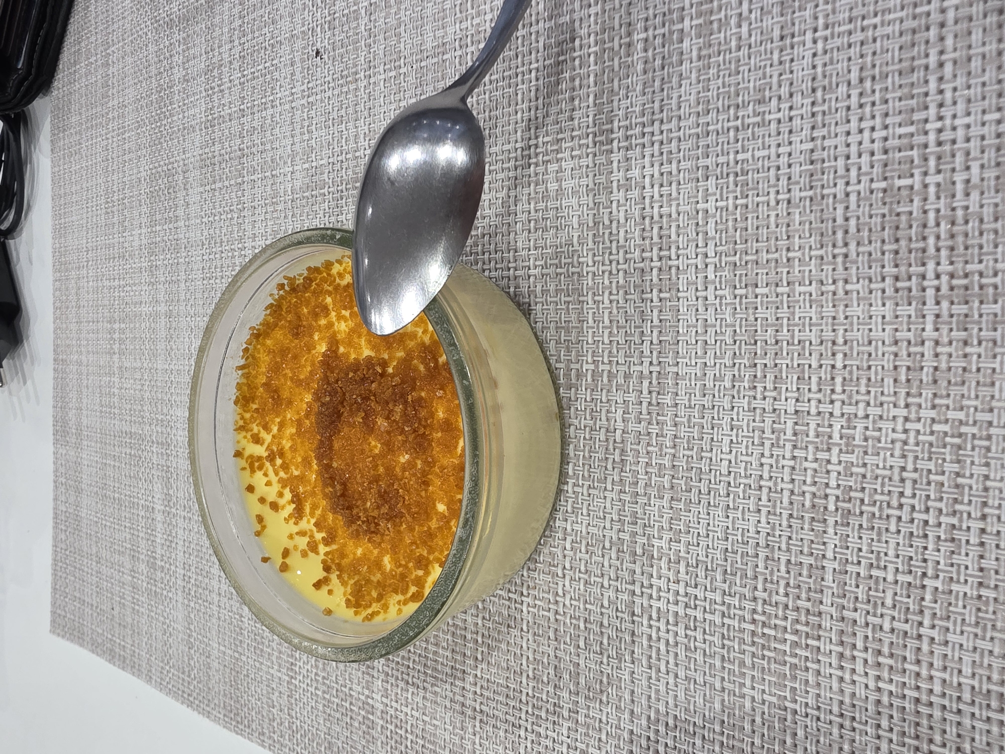 A glass bowl containing a yellow custard-like dessert with a caramelized sugar topping is positioned on a gray woven placemat. A silver spoon rests on the bowl. In the background, a portion of a black object and a white surface are visible. The location is not discernible from the image.