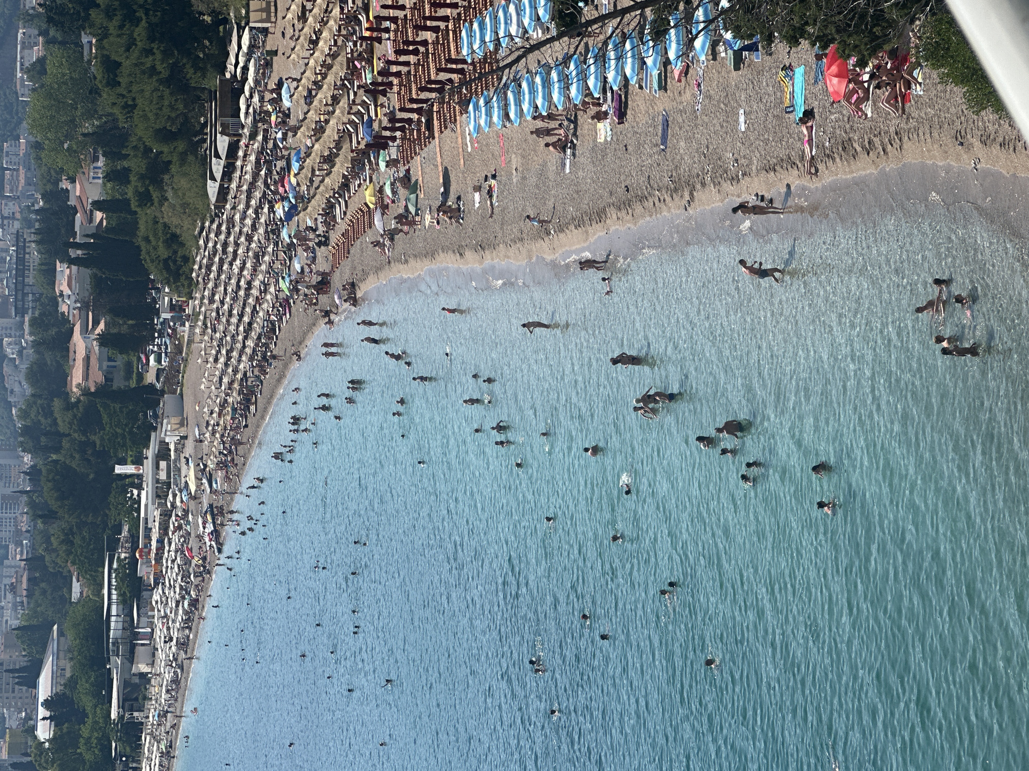 This image depicts a crowded beach scene in Budva, Montenegro, likely Mogren Beach, given its characteristic pebbly shore, clear blue water, and the backdrop of forested hills and urban development. The beach is lined with numerous rows of lounge chairs and umbrellas, creating a vibrant and bustling atmosphere. Many people are visible enjoying the water, swimming, and wading. Others are sunbathing on the beach, relaxing under the umbrellas, or strolling along the shoreline. The water is a stunning turquoise, reflecting the bright sunlight. The presence of many tourists and locals indicates a popular recreational area. The overall scene conveys a sense of summer leisure and enjoyment.